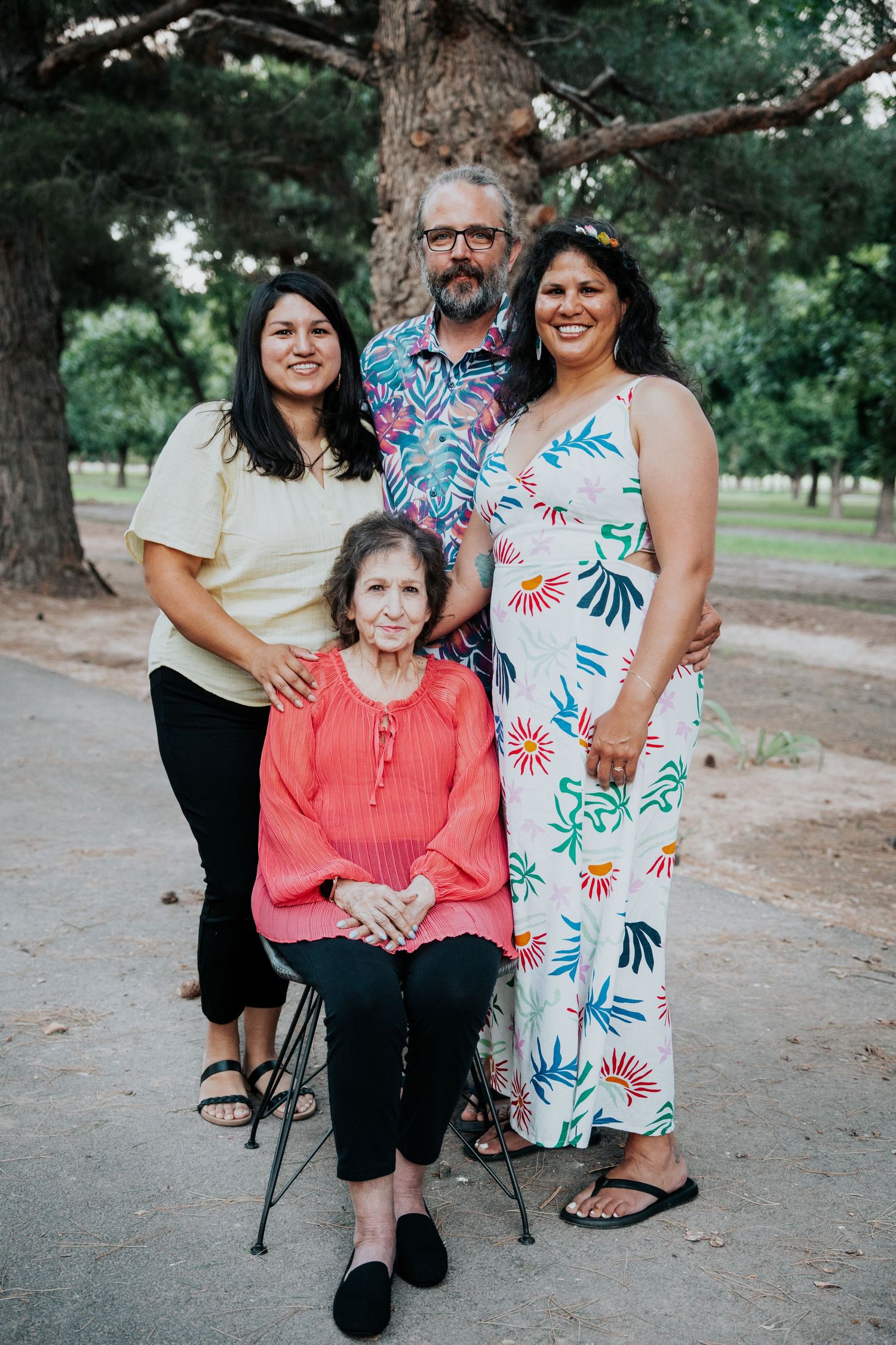 Family of four poses outdoors near a large tree, featuring two women, a bearded man, and an elderly woman seated.