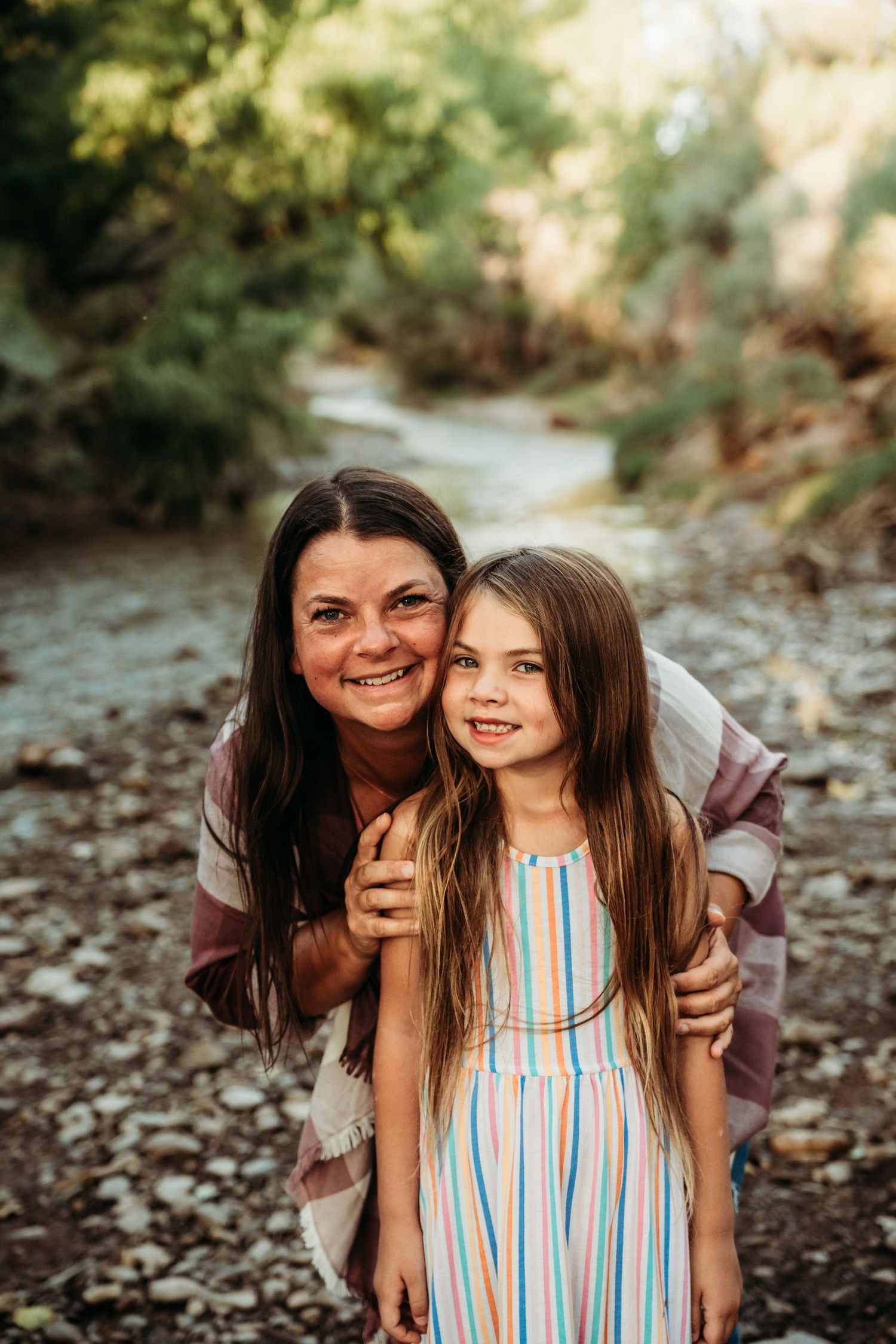 A smiling mother hugs her young daughter outdoors near a rocky creek surrounded by green trees.