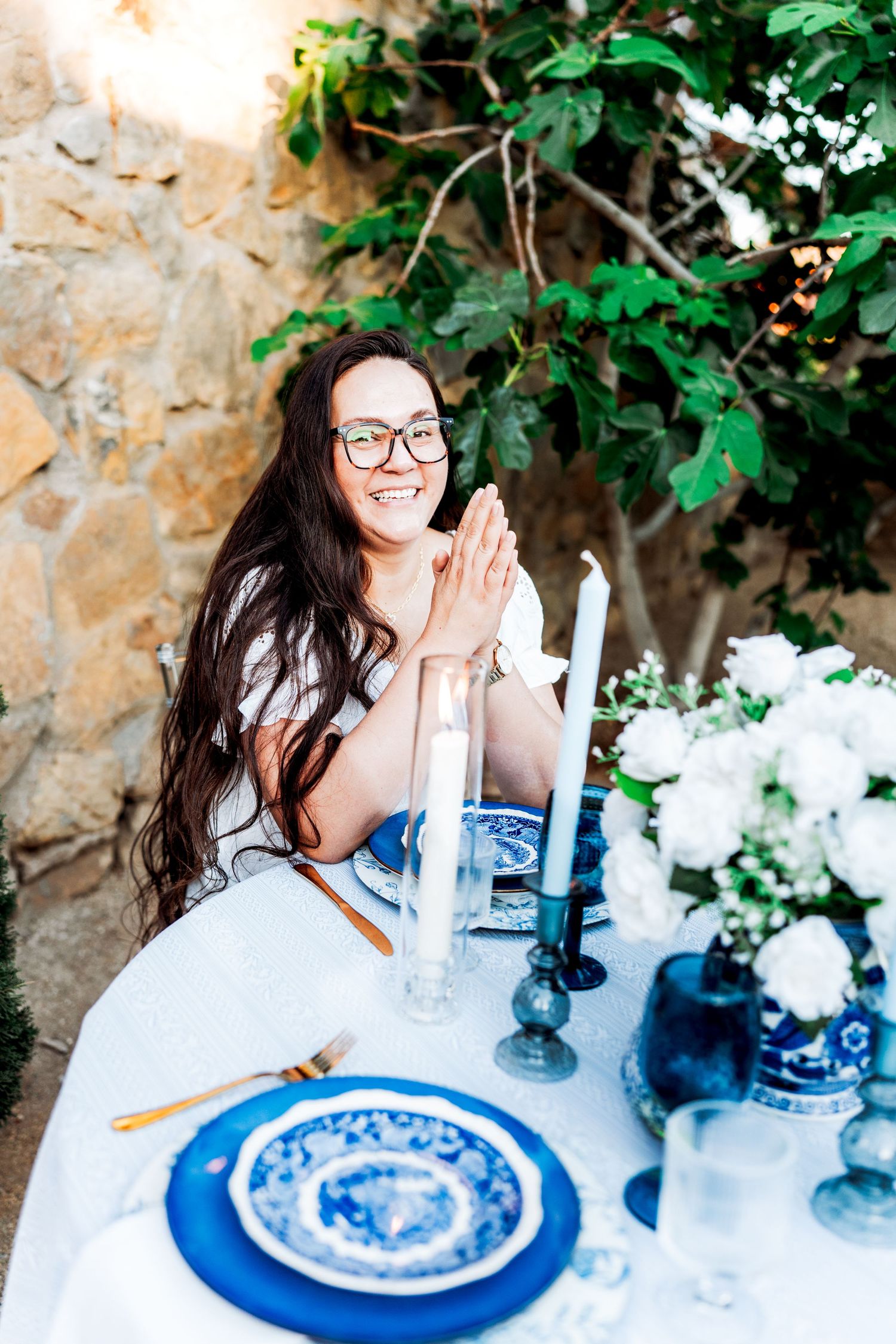 Smiling woman in white dress seated at elegant outdoor table with blue plates, white roses, and candles against stone wall.