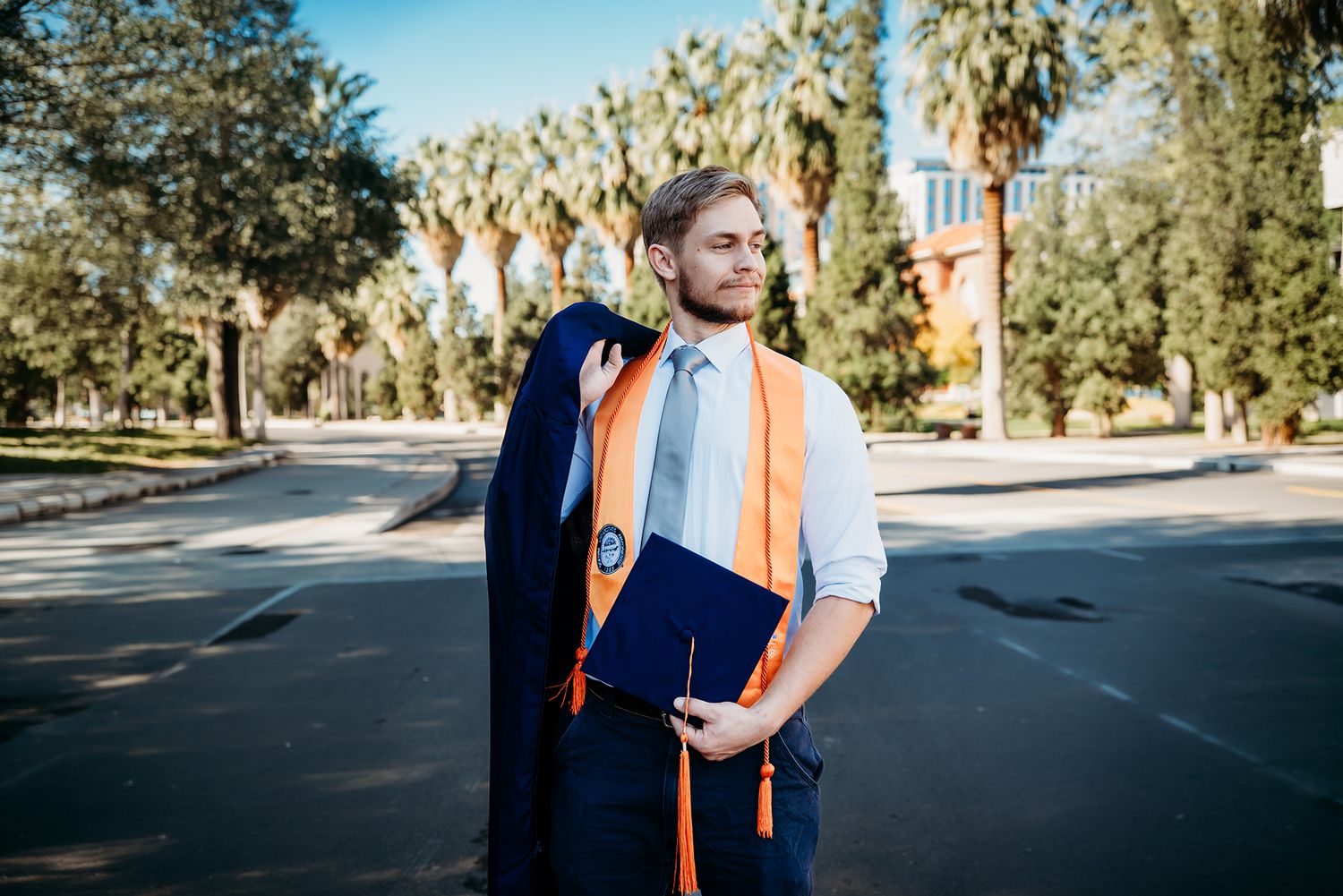 A young male graduate in orange and blue regalia holds a diploma outdoors near palm trees on a sunny day.