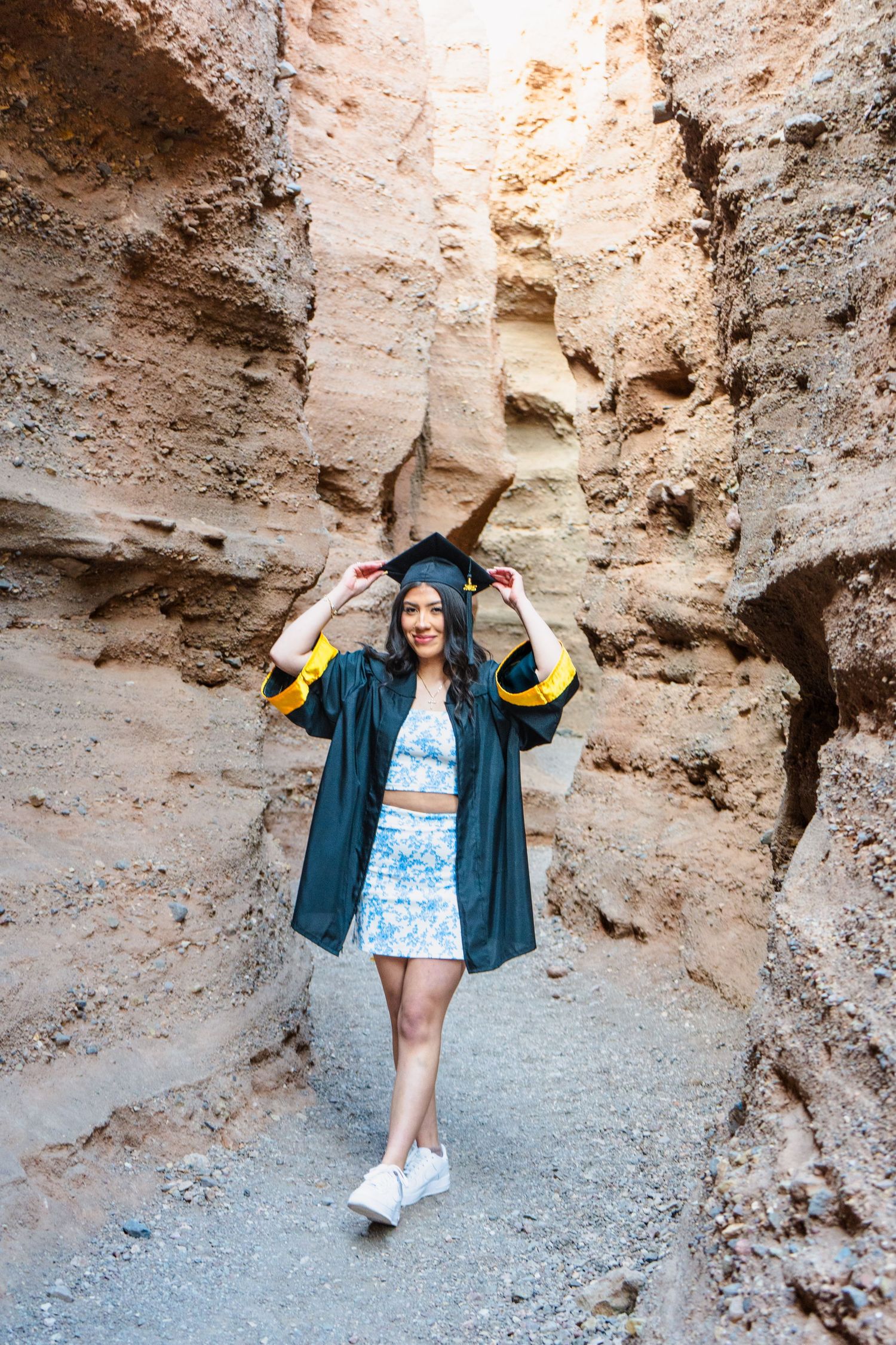 Female graduate in black and gold gown holding cap while walking through a sandstone canyon.