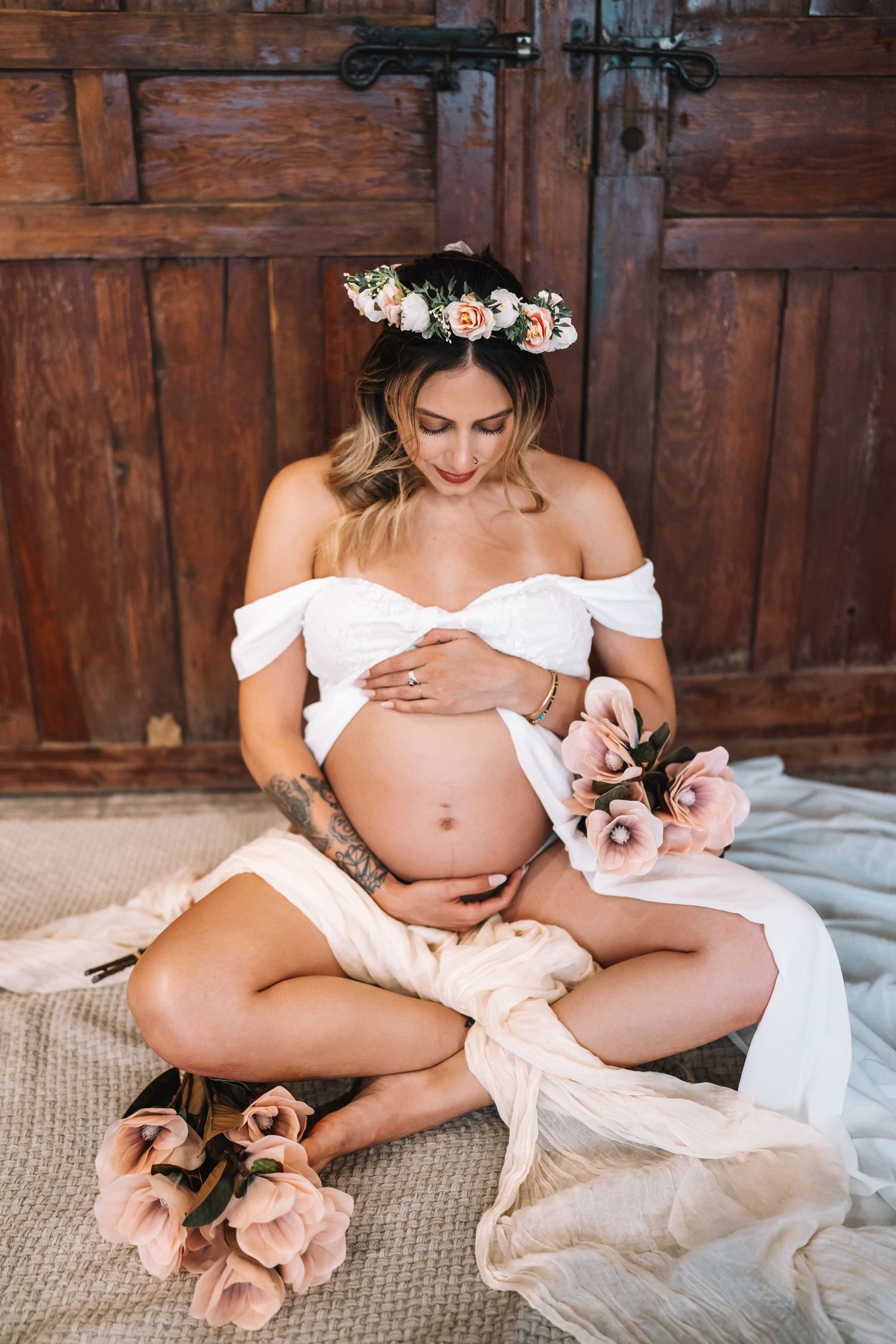 Pregnant woman wearing floral crown and white off-shoulder dress cradles baby bump surrounded by pink blooms.