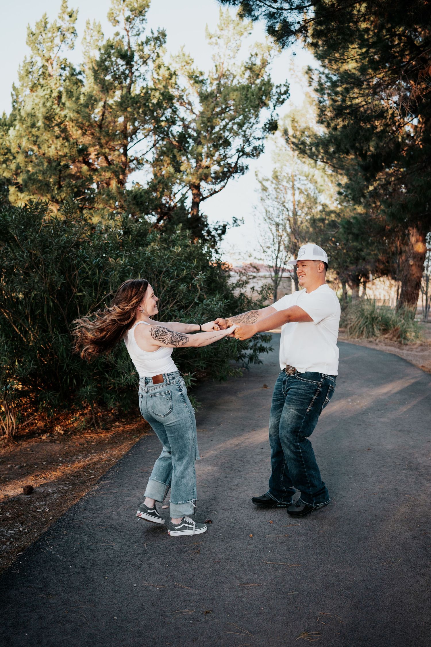 Joyful couple spinning and holding hands on a scenic tree-lined path at golden hour.