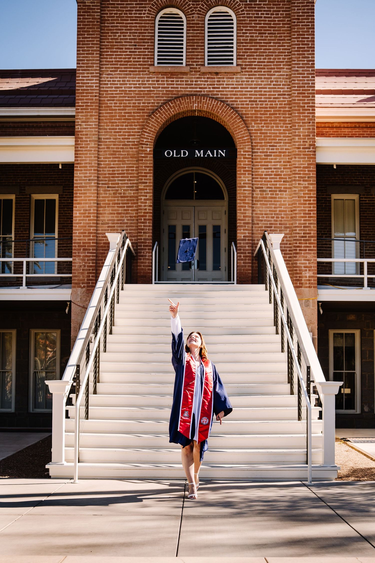 Graduate in cap and gown raises hand in front of Old Main brick university building.