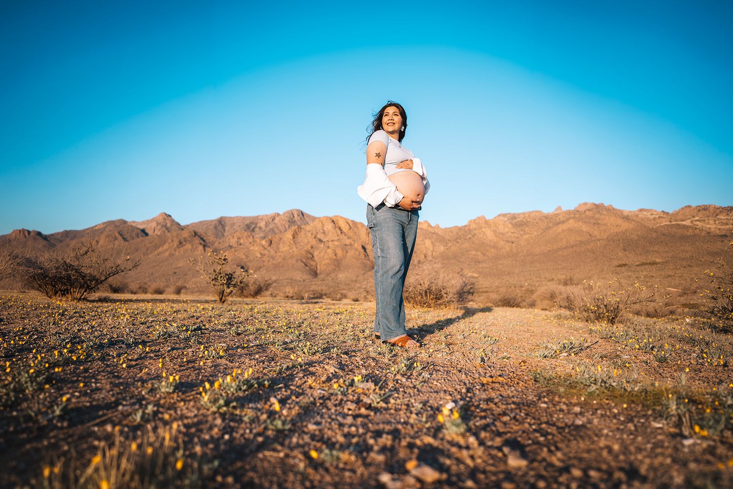 Pregnant woman walks joyfully through desert wildflower field with mountains in background.
