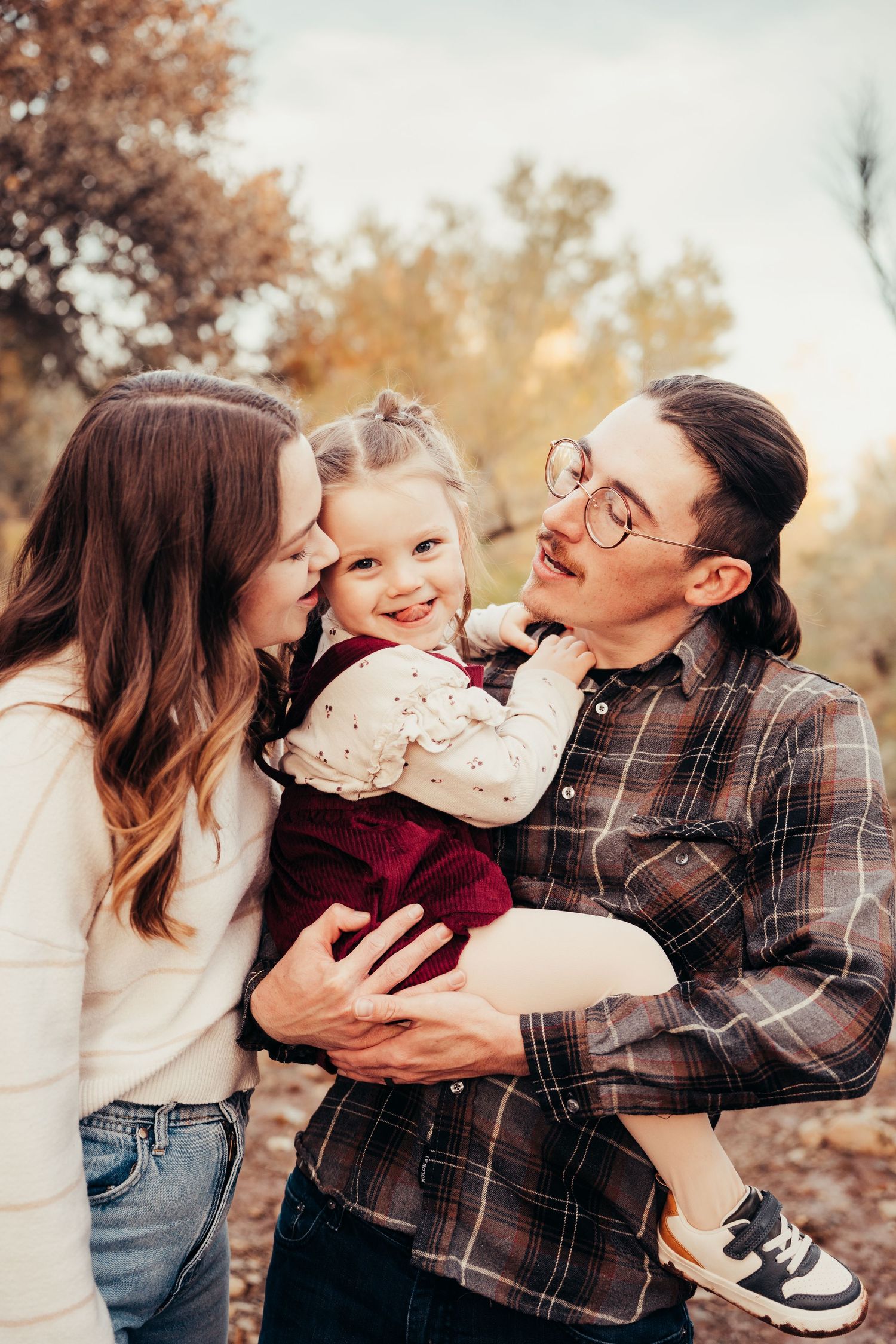 Happy young family with father holding smiling toddler girl as mother kisses her cheek in autumn park.