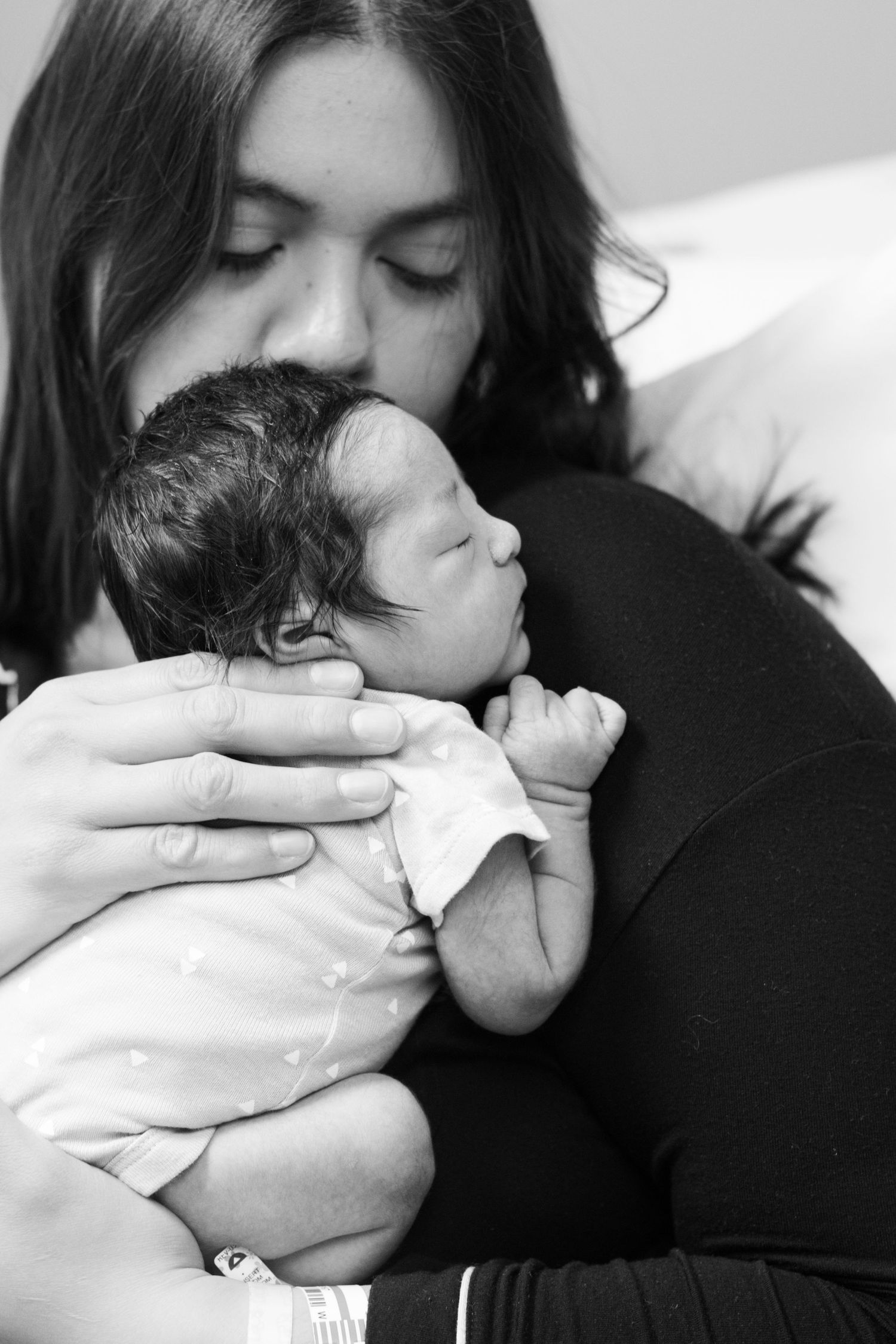 Black and white photo of mother kissing her newborn baby held close to her chest.
