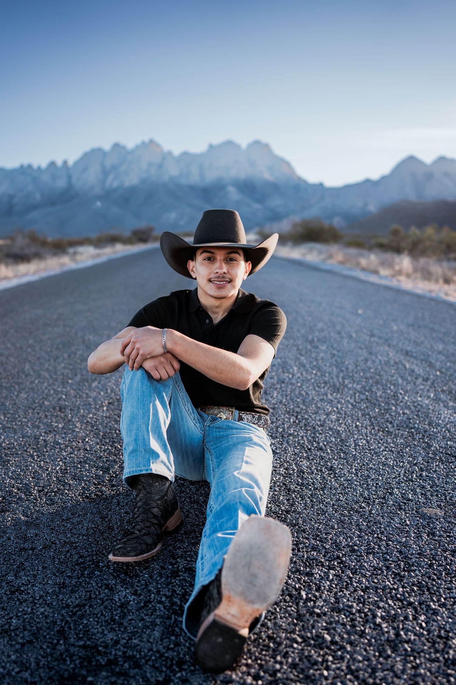 Young man in cowboy hat and jeans sitting relaxed on open road with mountains behind.