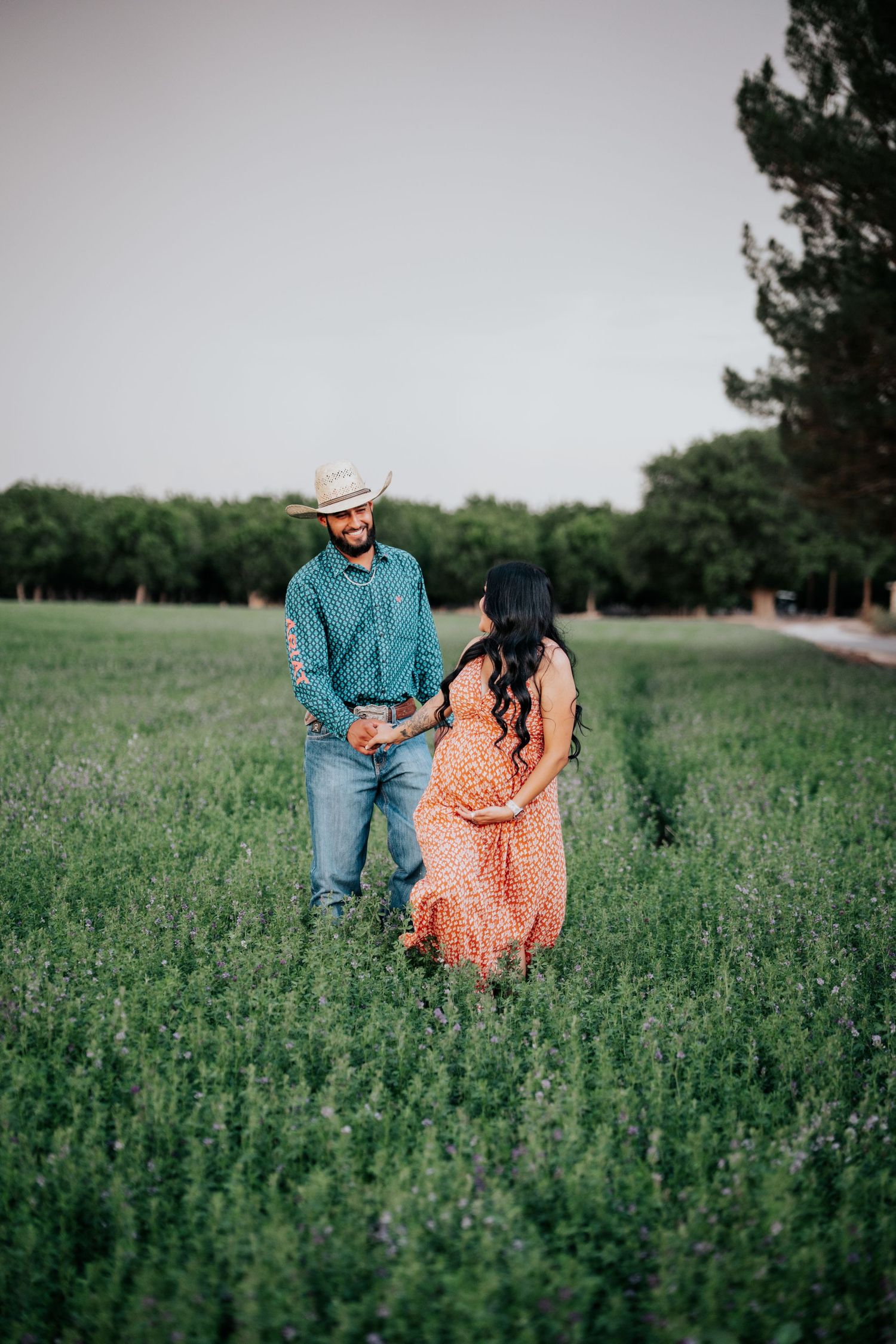 Couple holding hands in a green field, pregnant woman in orange floral dress facing man in cowboy hat and teal shirt.