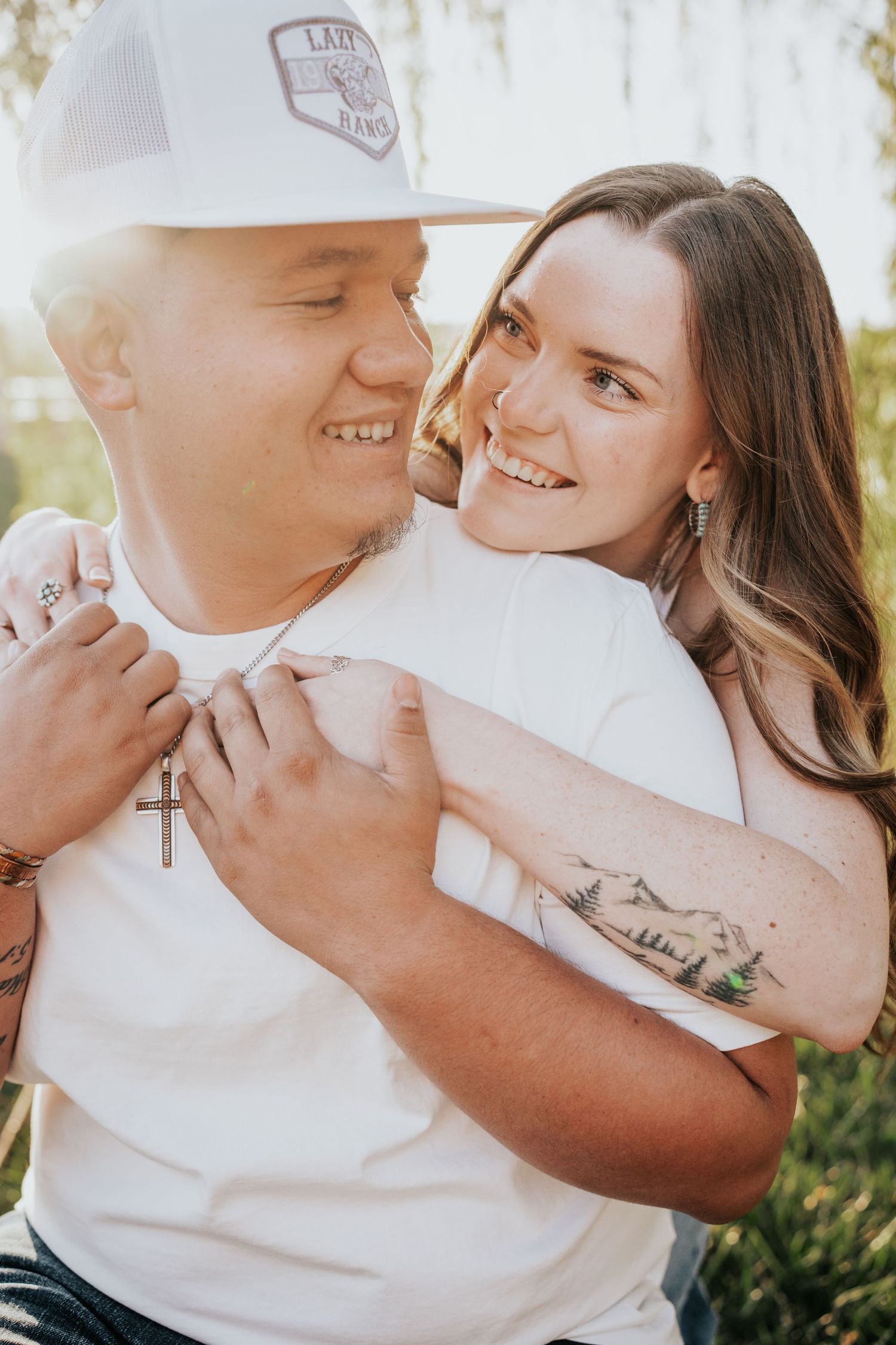 Young couple embracing outdoors, woman with mountain tattoo hugging smiling man in white cap and cross necklace.