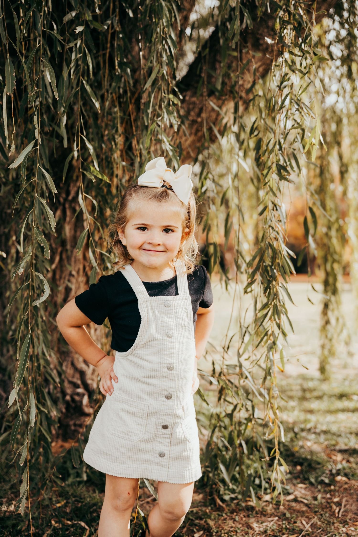 Adorable toddler girl in white overalls and bow smiling under a weeping willow tree outdoors.