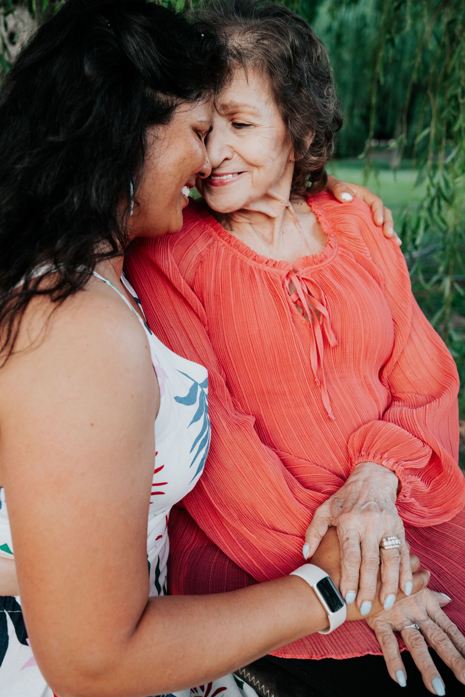 Two women sharing a tender forehead-to-forehead moment outdoors, one wearing coral blouse and a ring.