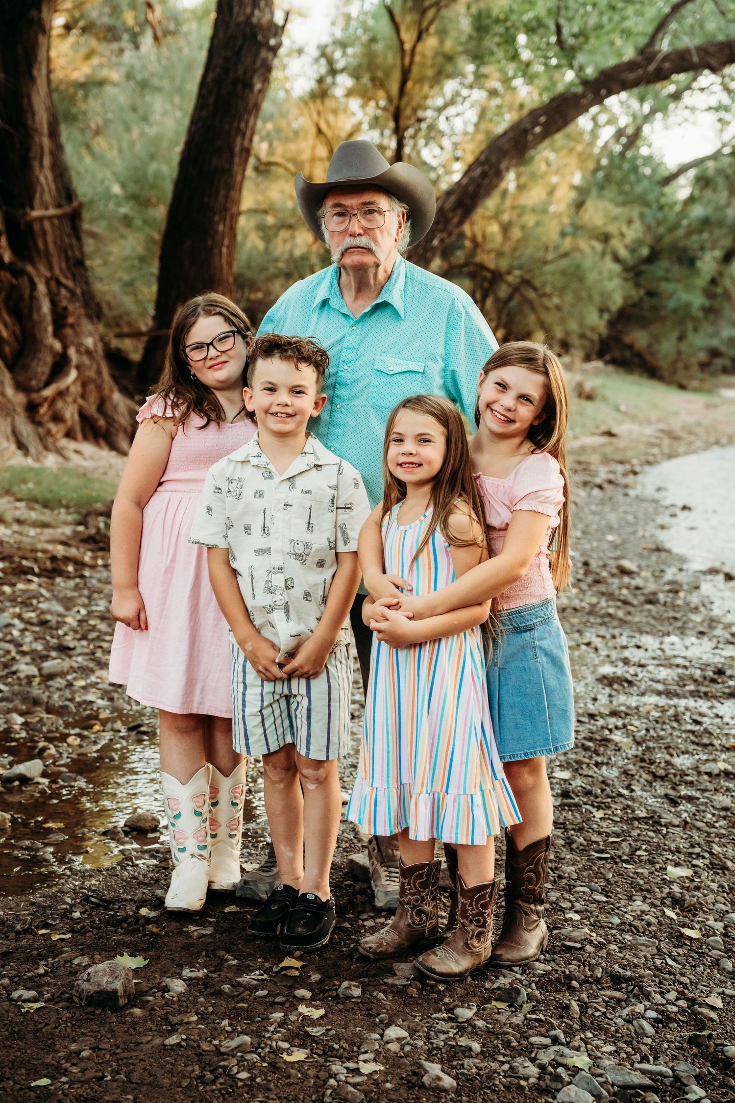 Grandfather in cowboy hat poses with four grandchildren by a rocky creek.