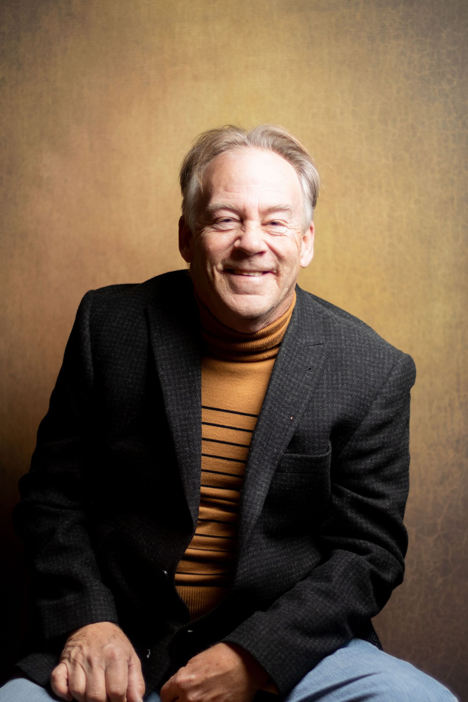 Smiling older man in black blazer and striped turtleneck seated casually.