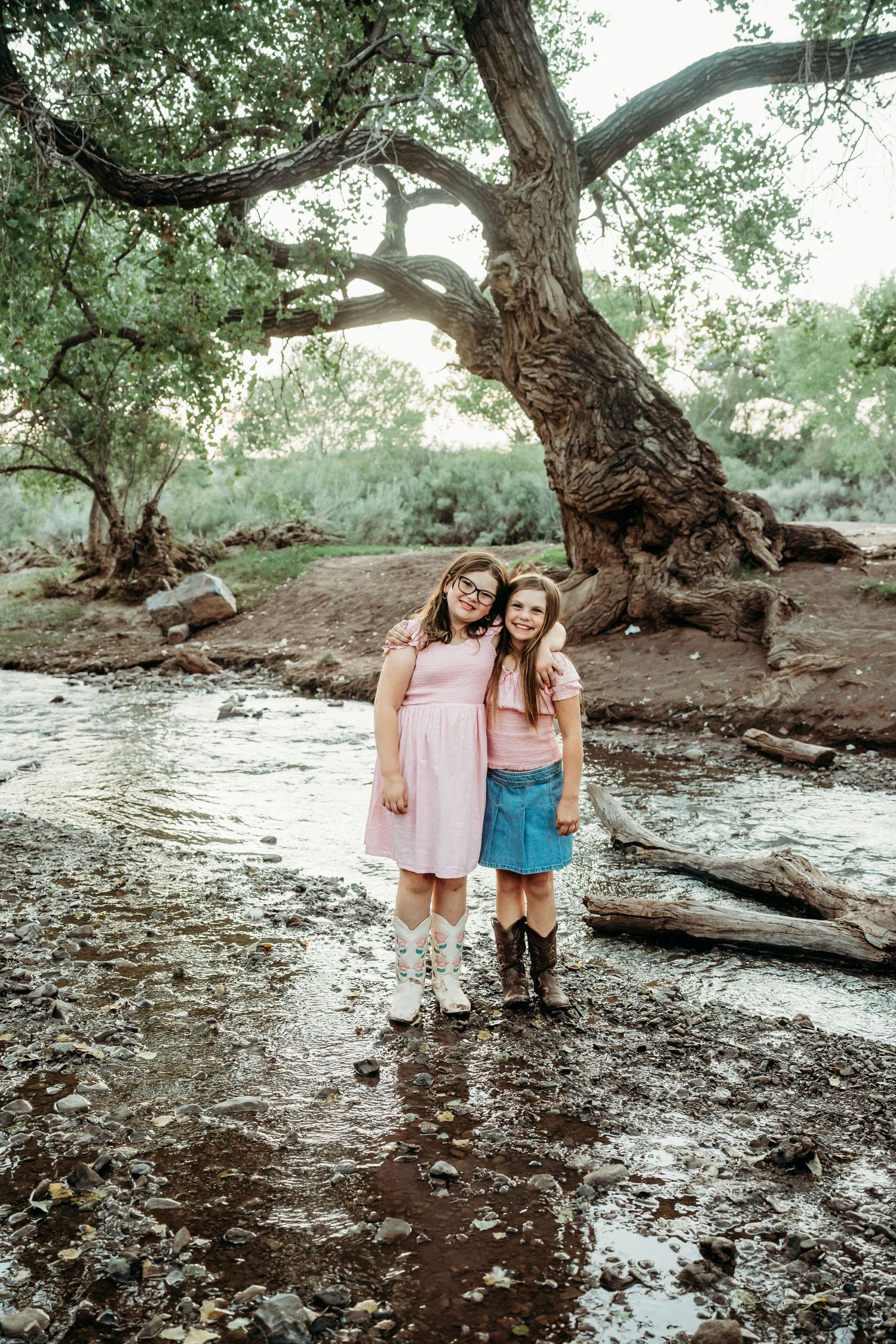 Two girls in matching pink tops smiling together by a river beneath a large tree.