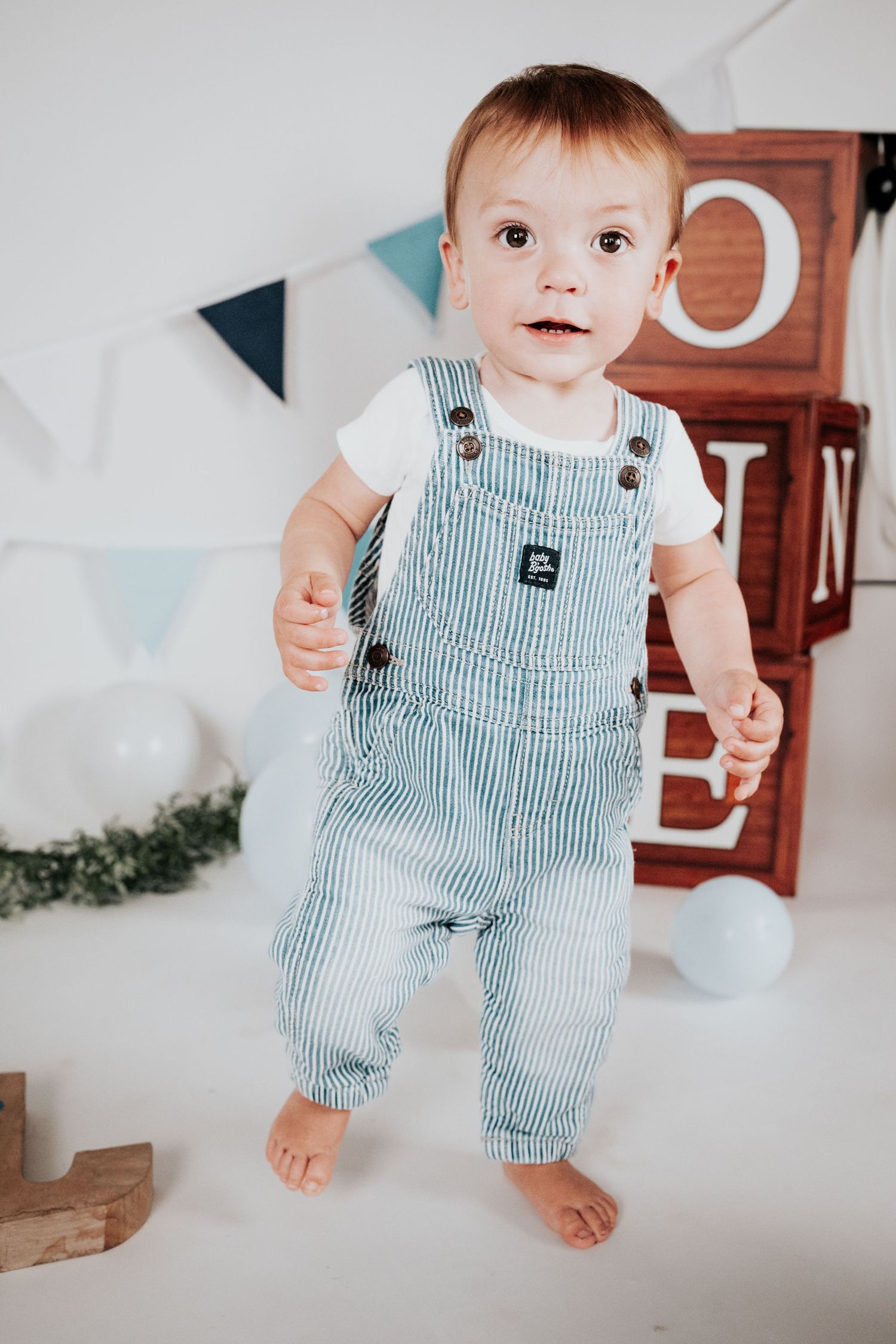Baby boy in striped overalls standing before ONE letter blocks at first birthday photoshoot.