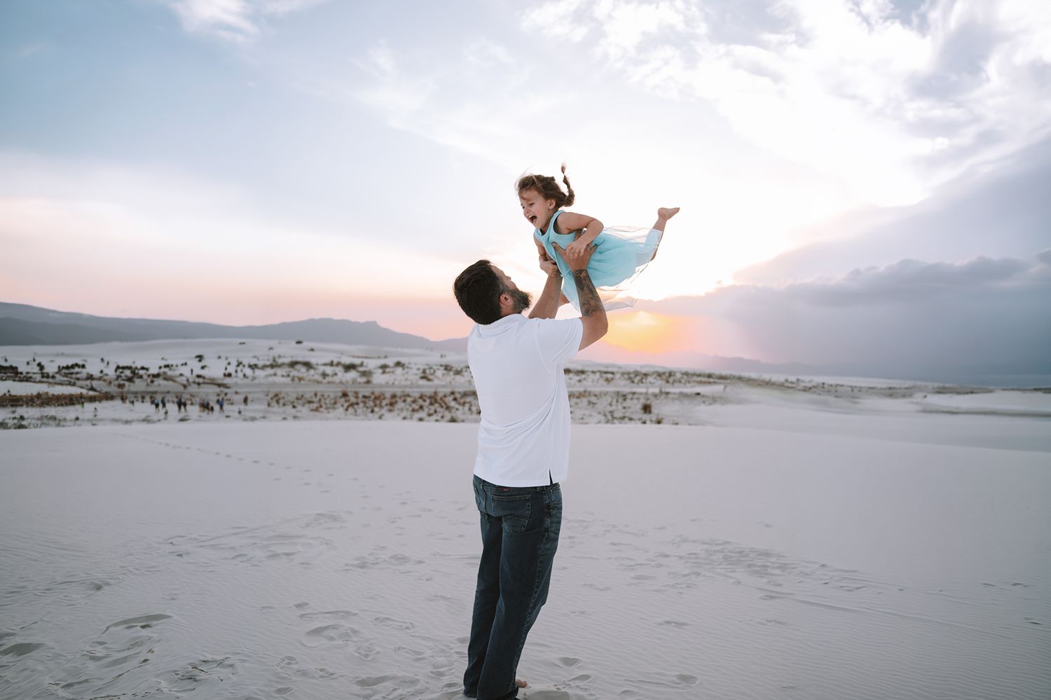 A father joyfully lifts his young daughter into the air on a sandy beach at sunset.