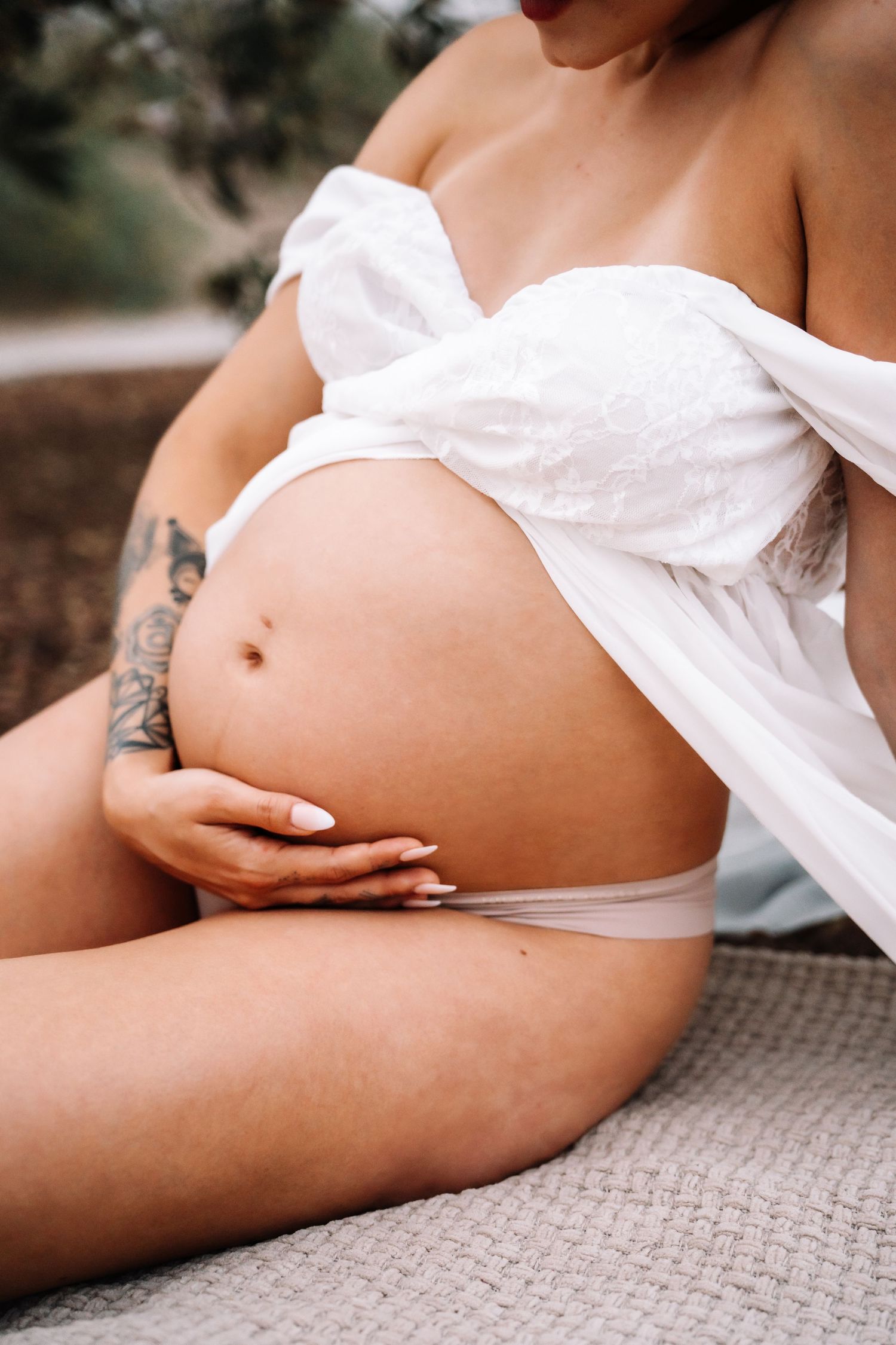 Close-up of pregnant woman in white off-shoulder top with tattoo cradling bare bump while seated outdoors.