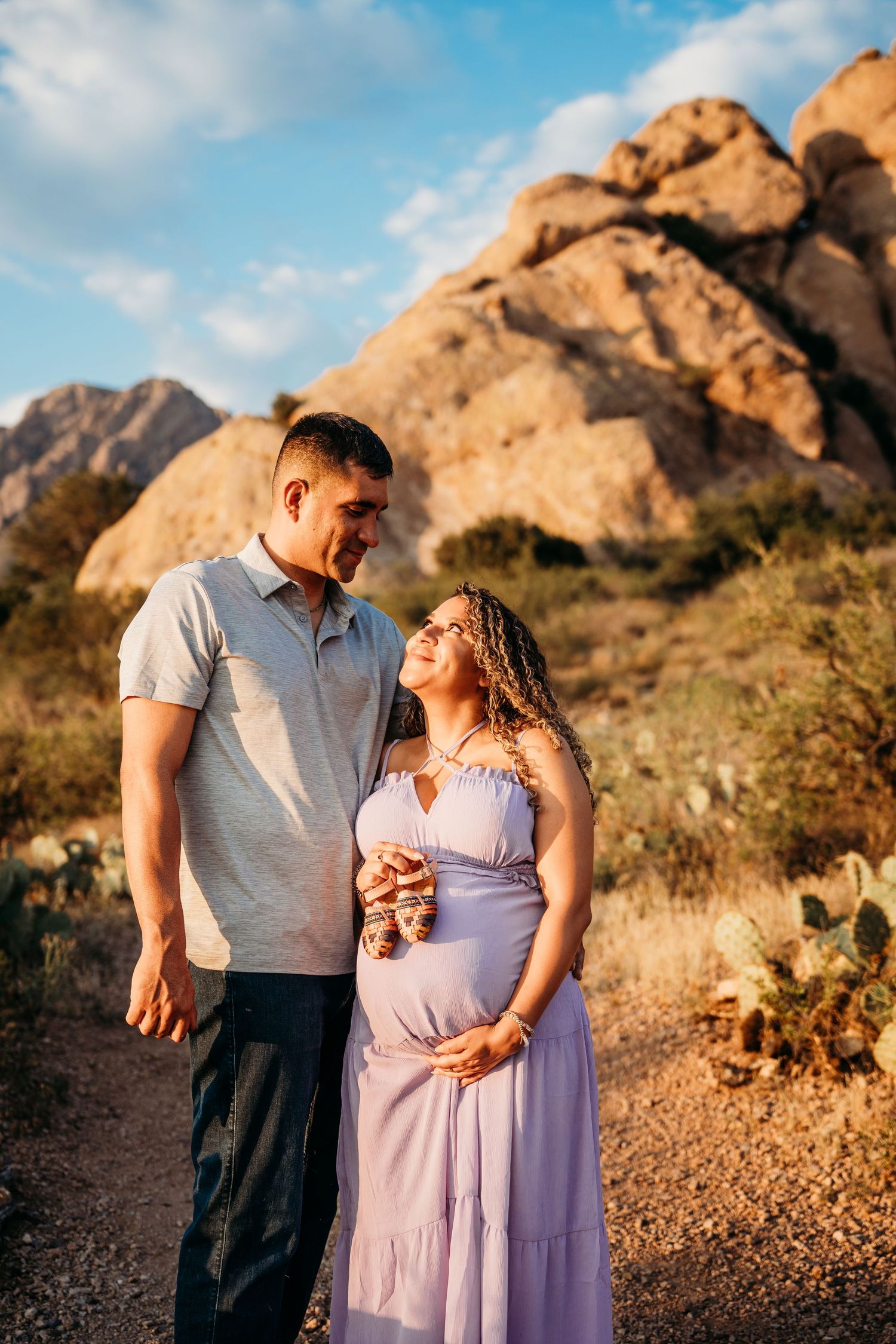 Pregnant couple gazing at each other in golden desert light, woman in purple dress holding baby booties, rocks behind.