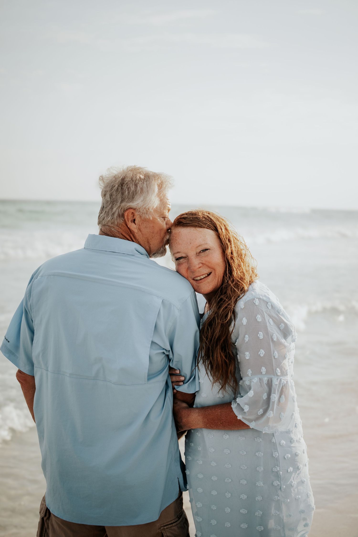Man kissing woman on cheek at beach, woman smiling joyfully, both wearing light blue outfits by ocean waves.