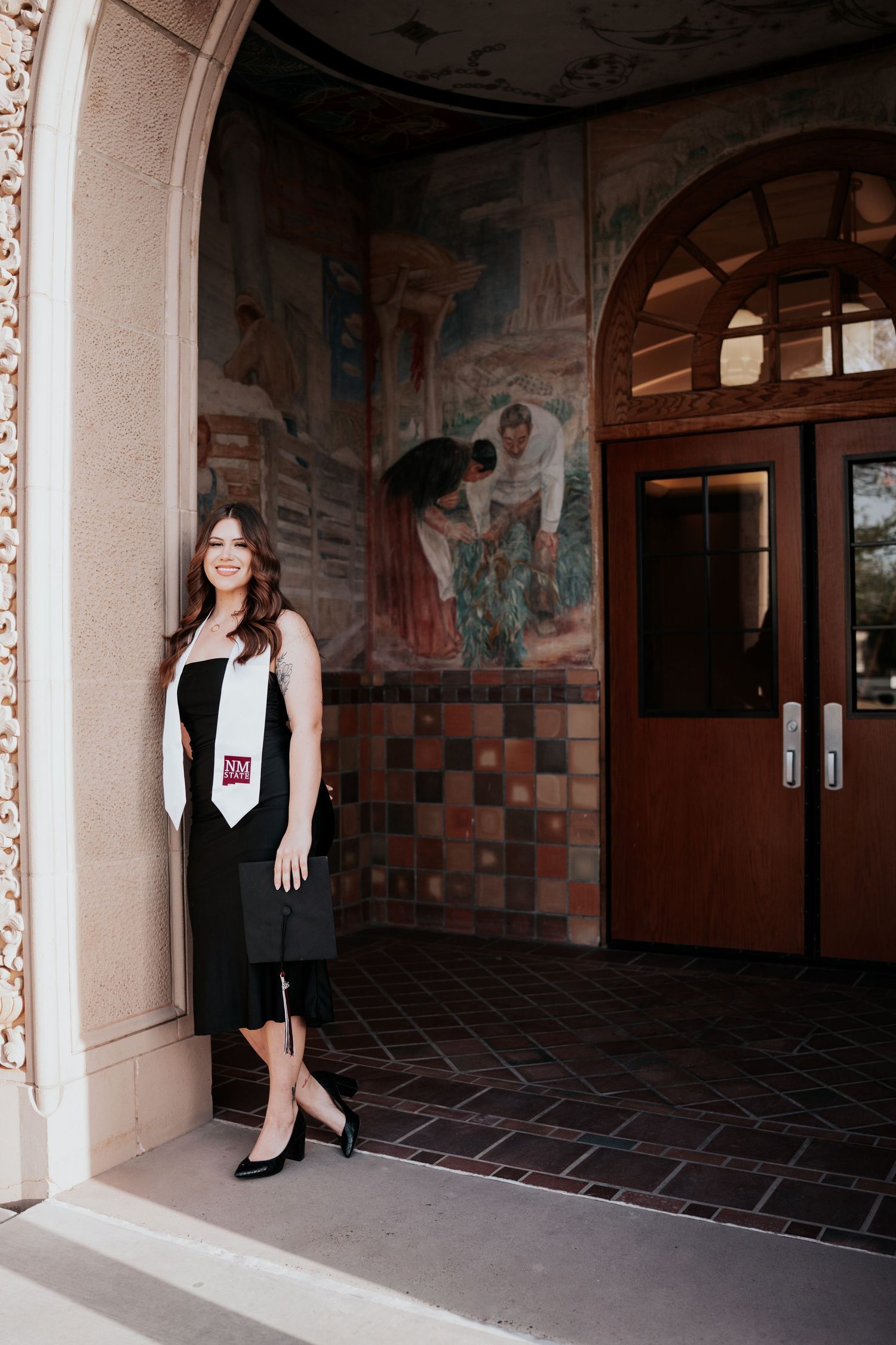 Female graduate in black cap and gown with NM State stole posing at ornate building entrance.