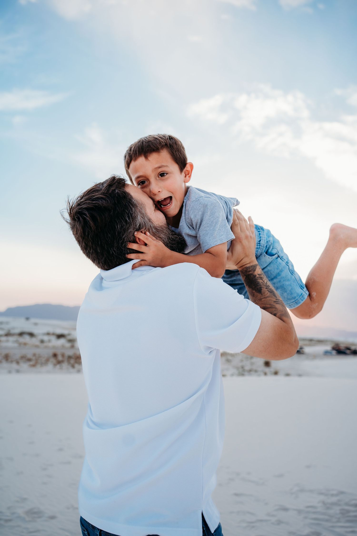A father lifts and kisses his excited young son against a blue sky on white sand dunes.