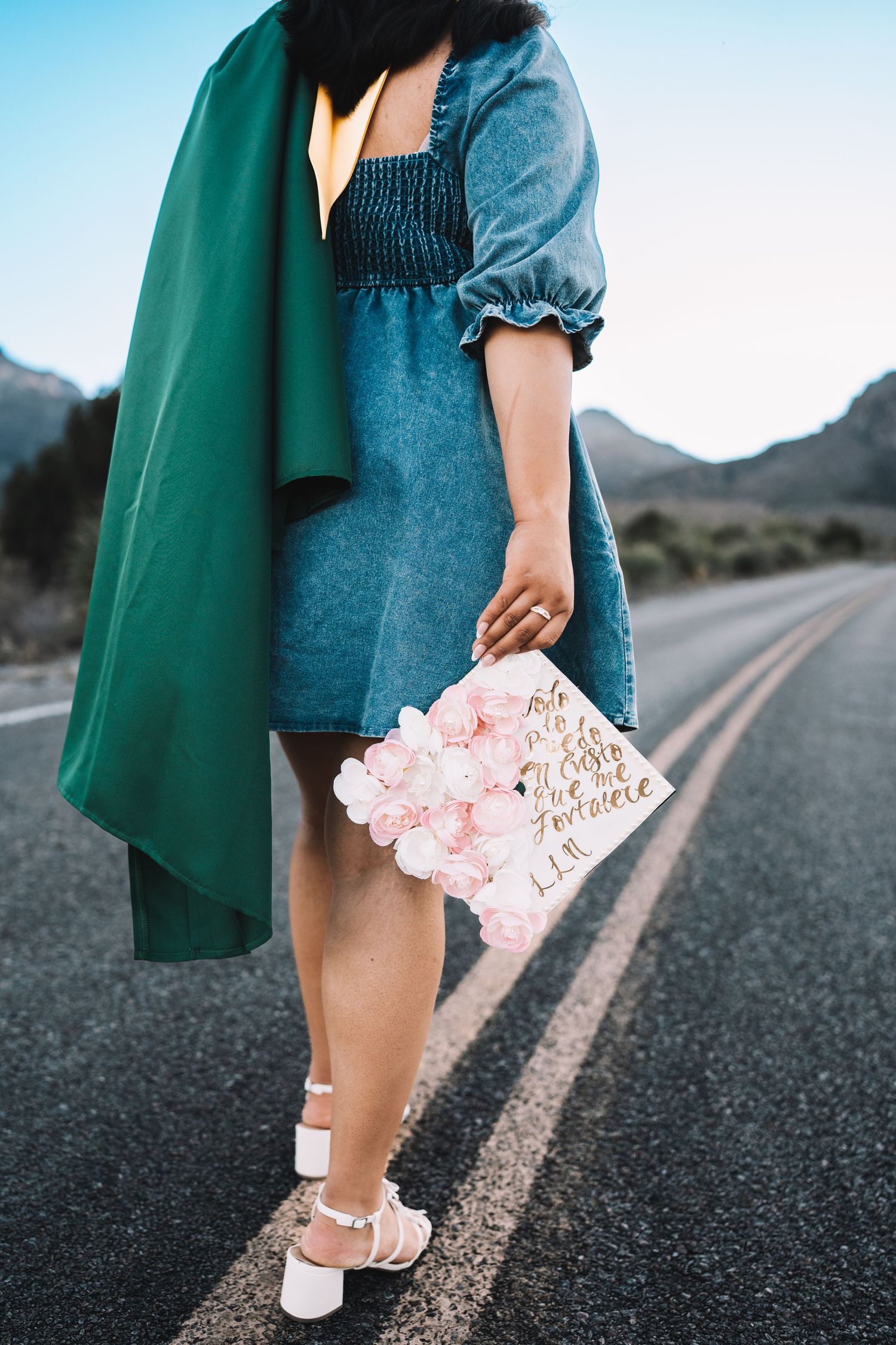 Close-up of a graduate in a denim dress holding a flower-decorated graduation cap on a desert highway.