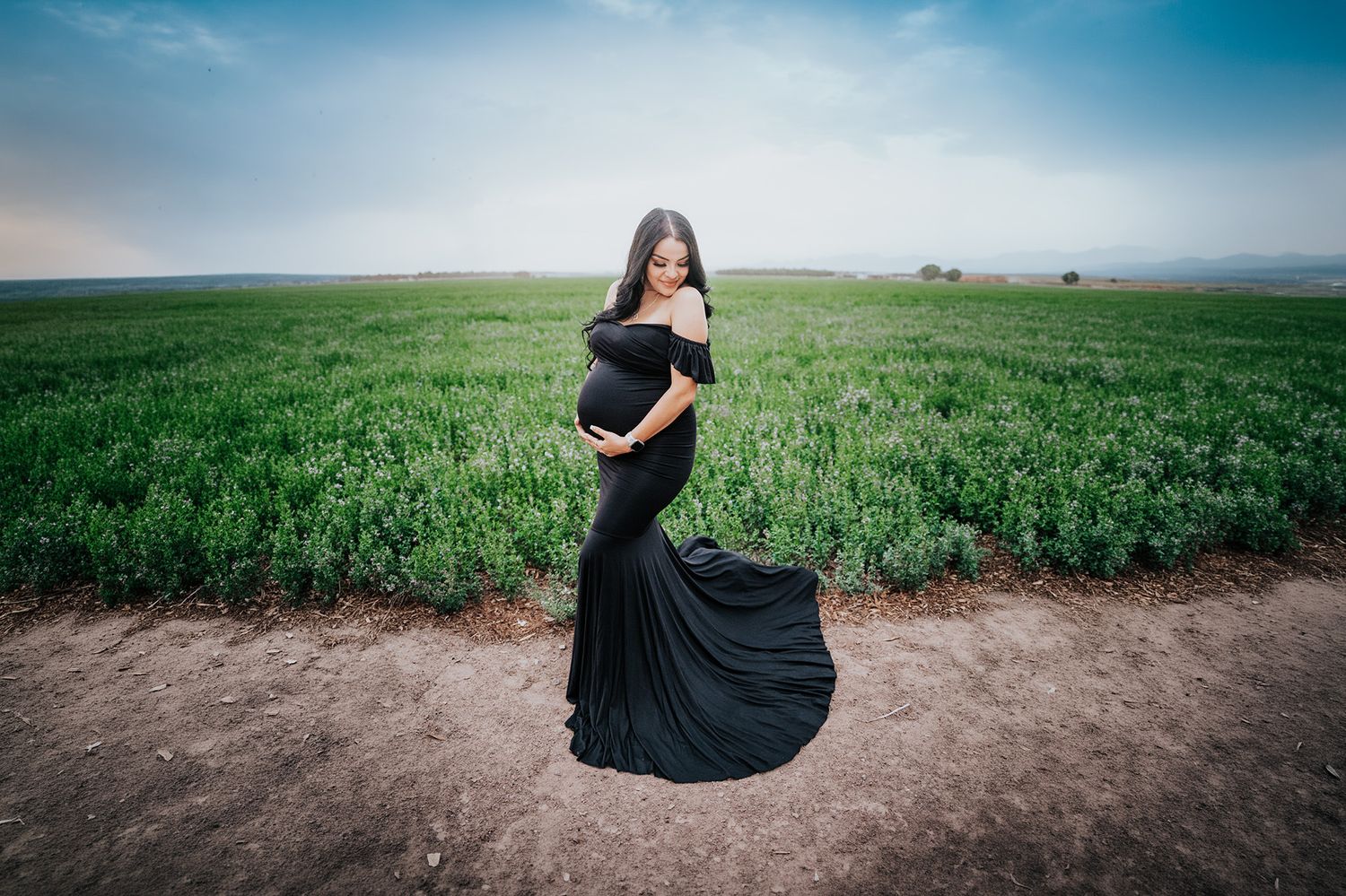 Elegant pregnant woman in a flowing black mermaid dress standing in a lush green field.