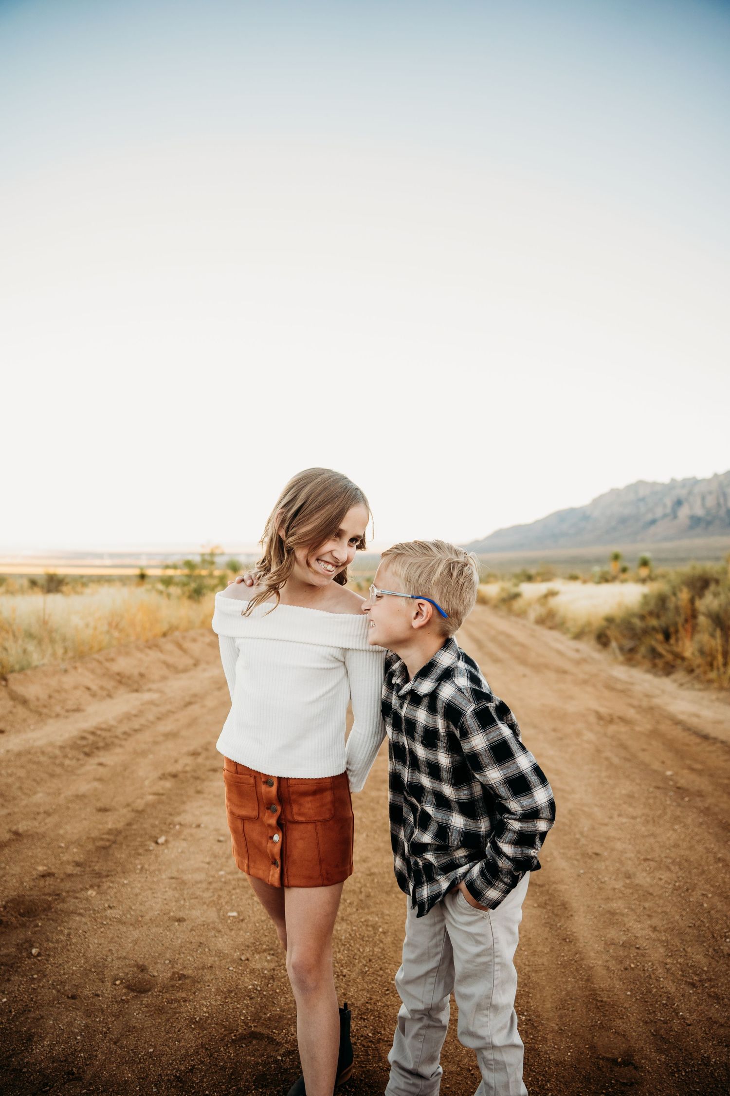A girl in a white top and rust skirt shares a sweet moment with a boy outdoors.