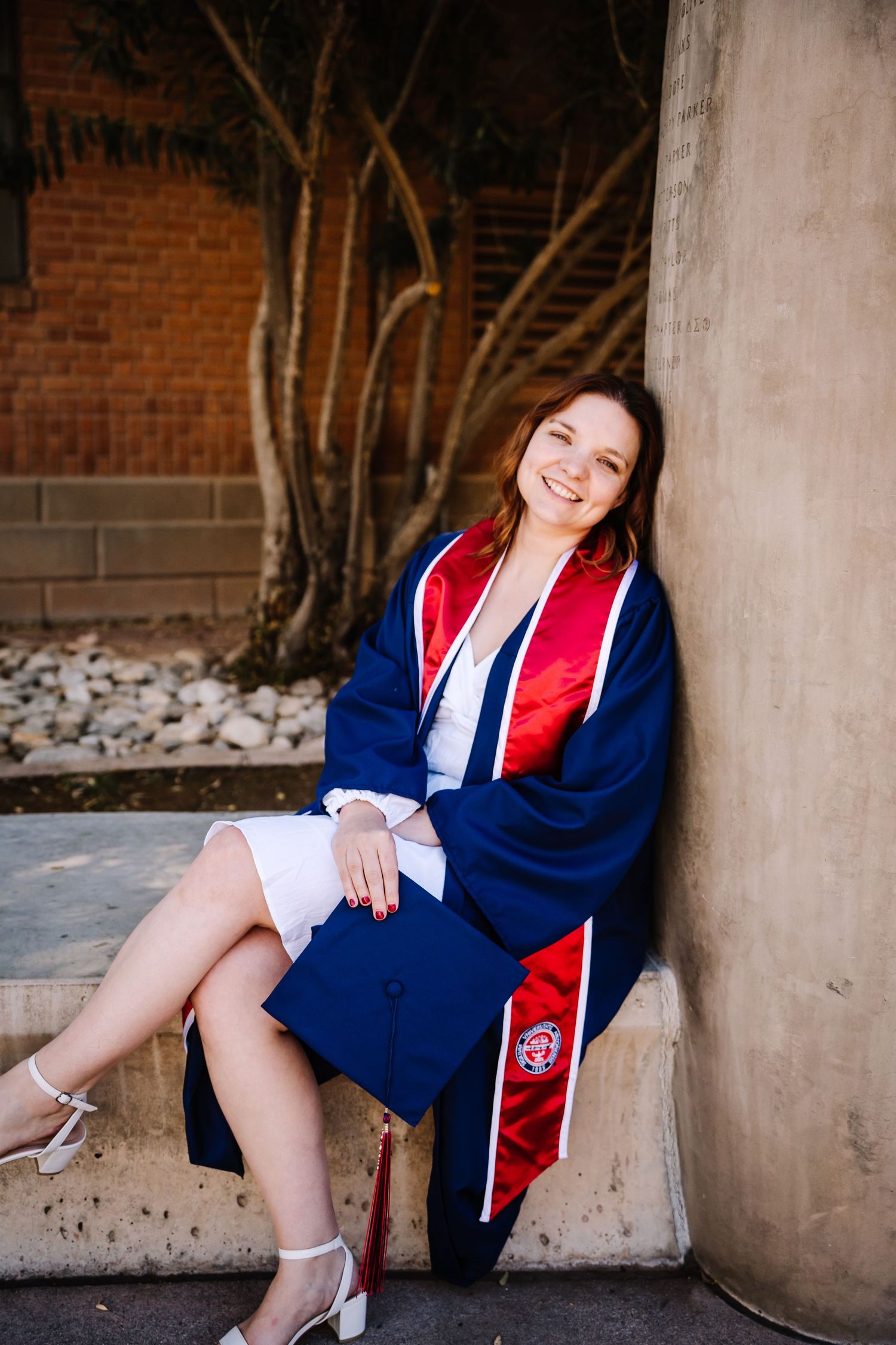 Smiling young woman in navy blue graduation gown with red stole sits outdoors leaning against a concrete pillar.