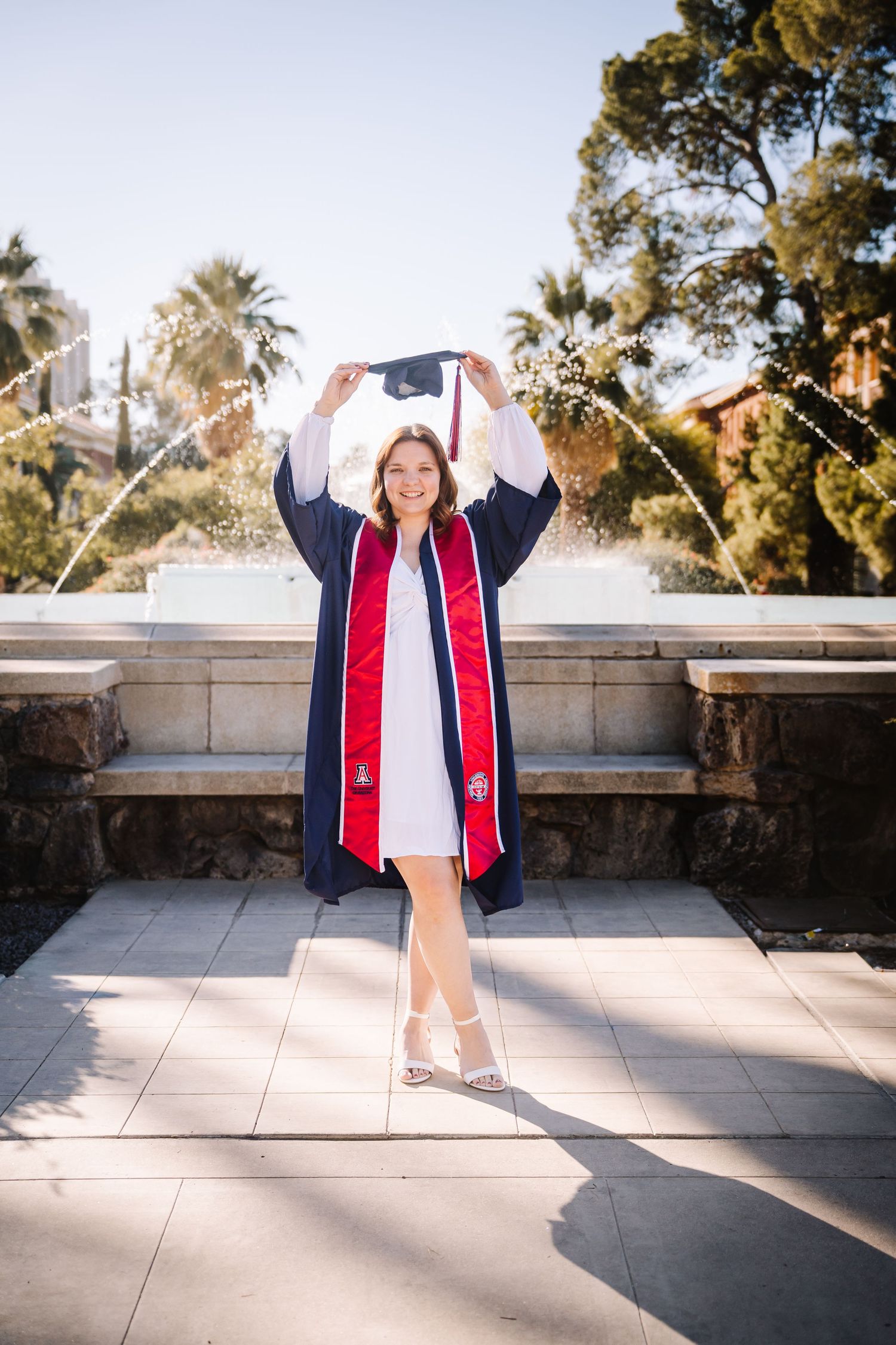 Graduate in navy cap and gown with red stole holds cap overhead near fountain, celebrating college graduation outdoors.