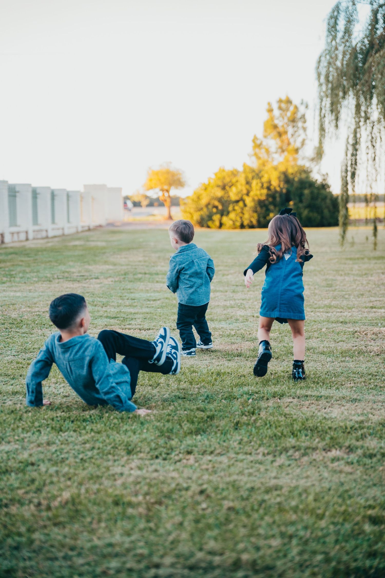 Three young children playing freely on a grassy park lawn during a warm golden sunset afternoon.