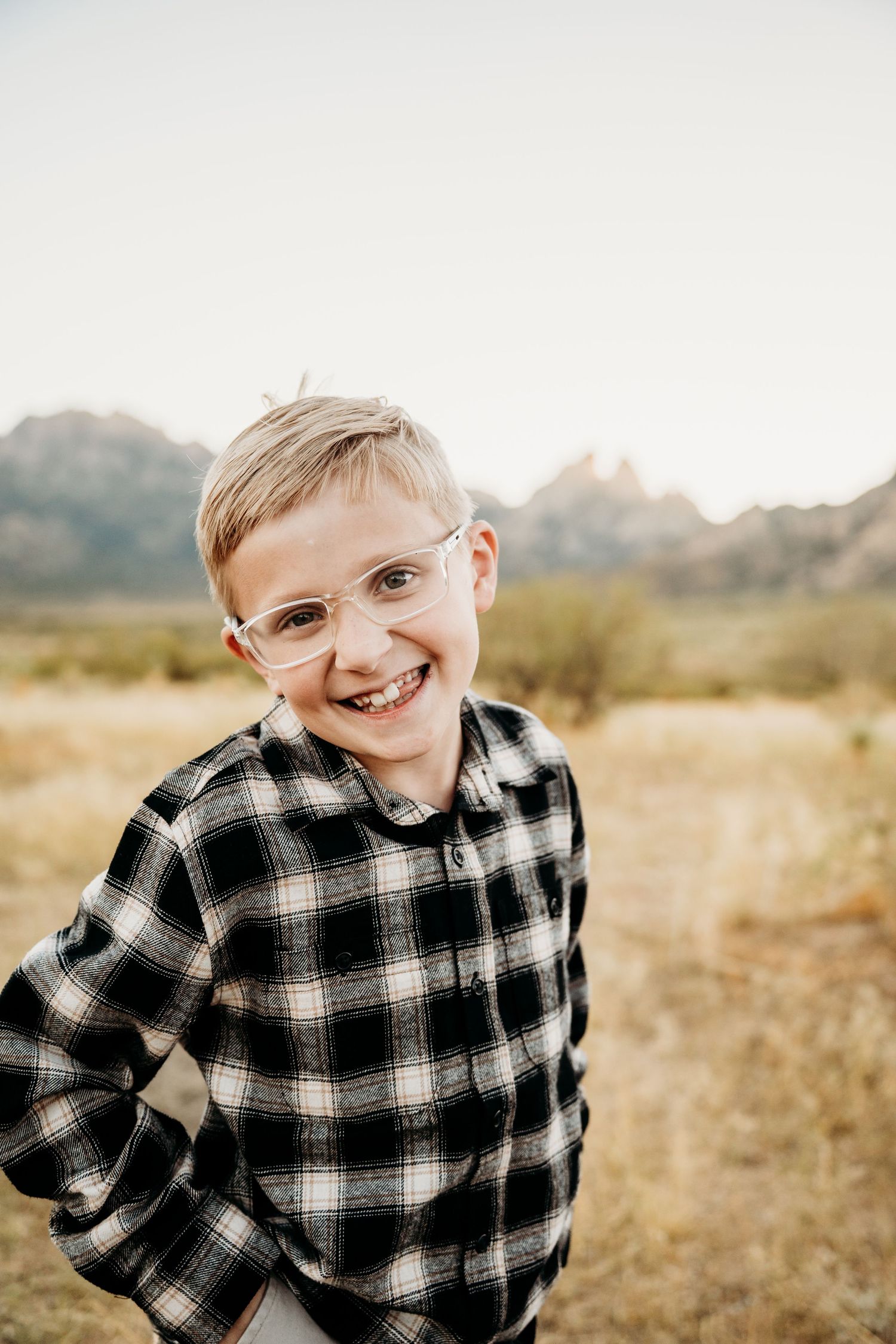Cheerful young boy in plaid shirt and glasses smiles confidently in a desert mountain setting.