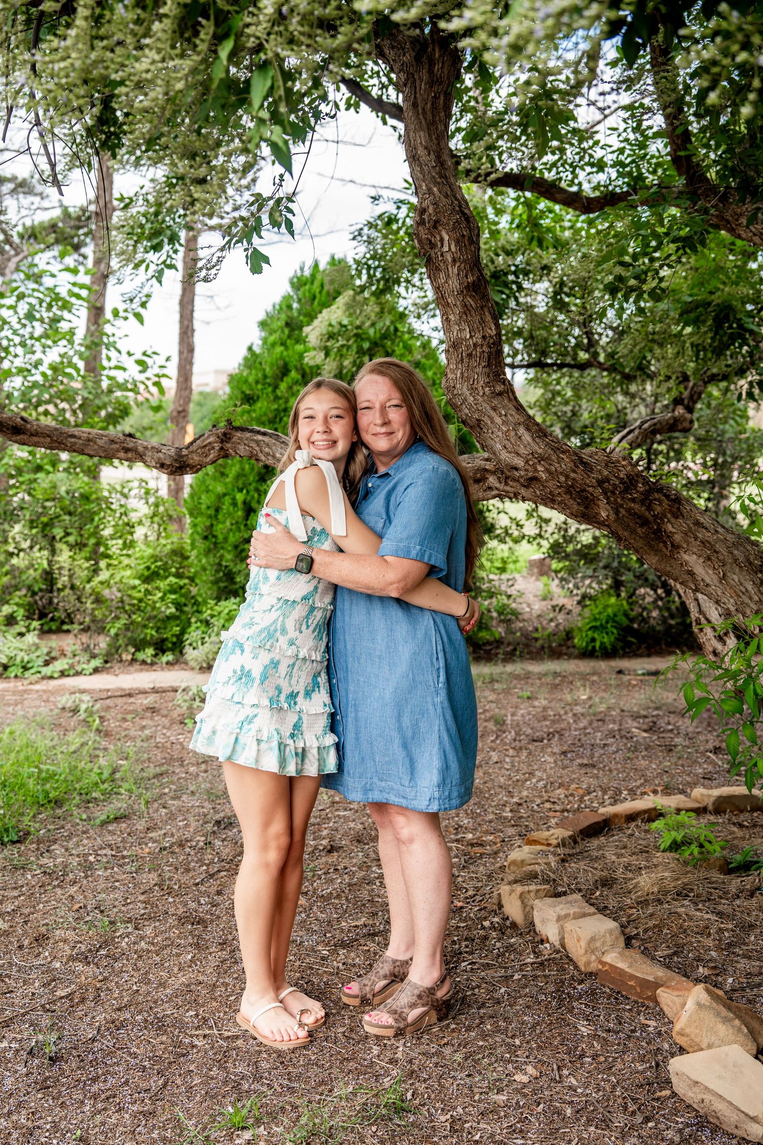Two girls in summer dresses share a sweet hug beneath a sprawling tree in a garden.