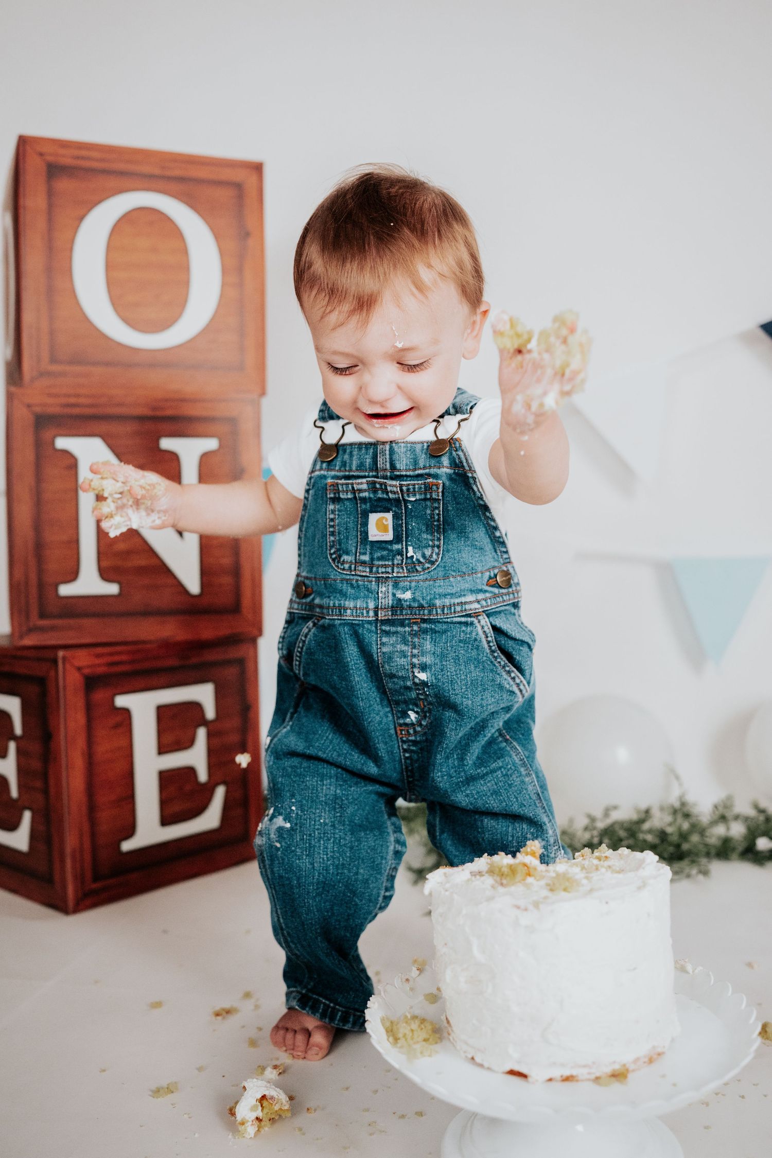 Joyful toddler in denim overalls standing and celebrating with cake-covered hands at first birthday party.