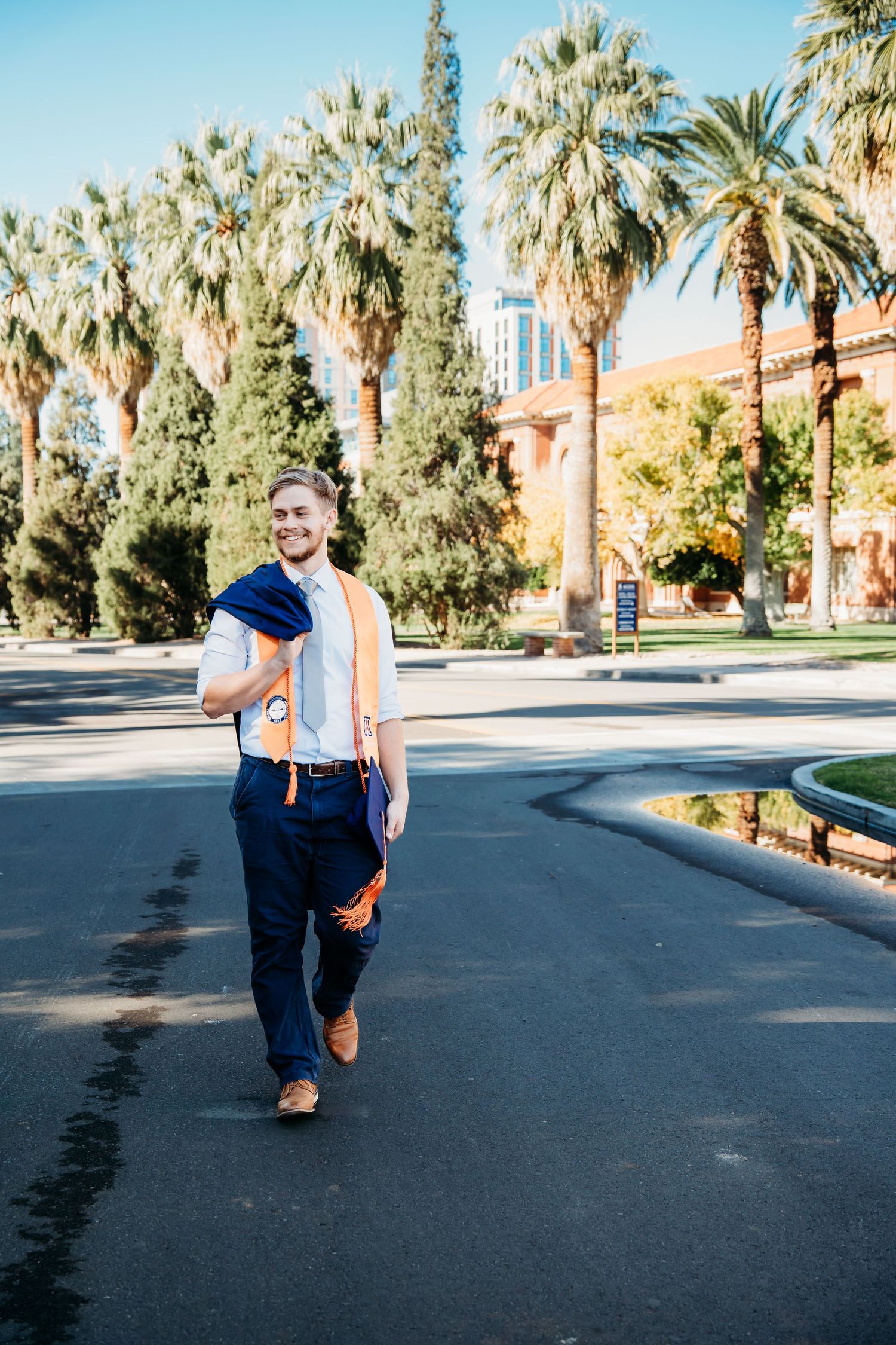 Smiling male college graduate walks outdoors in blue pants and orange sash among palm trees on a sunny campus.