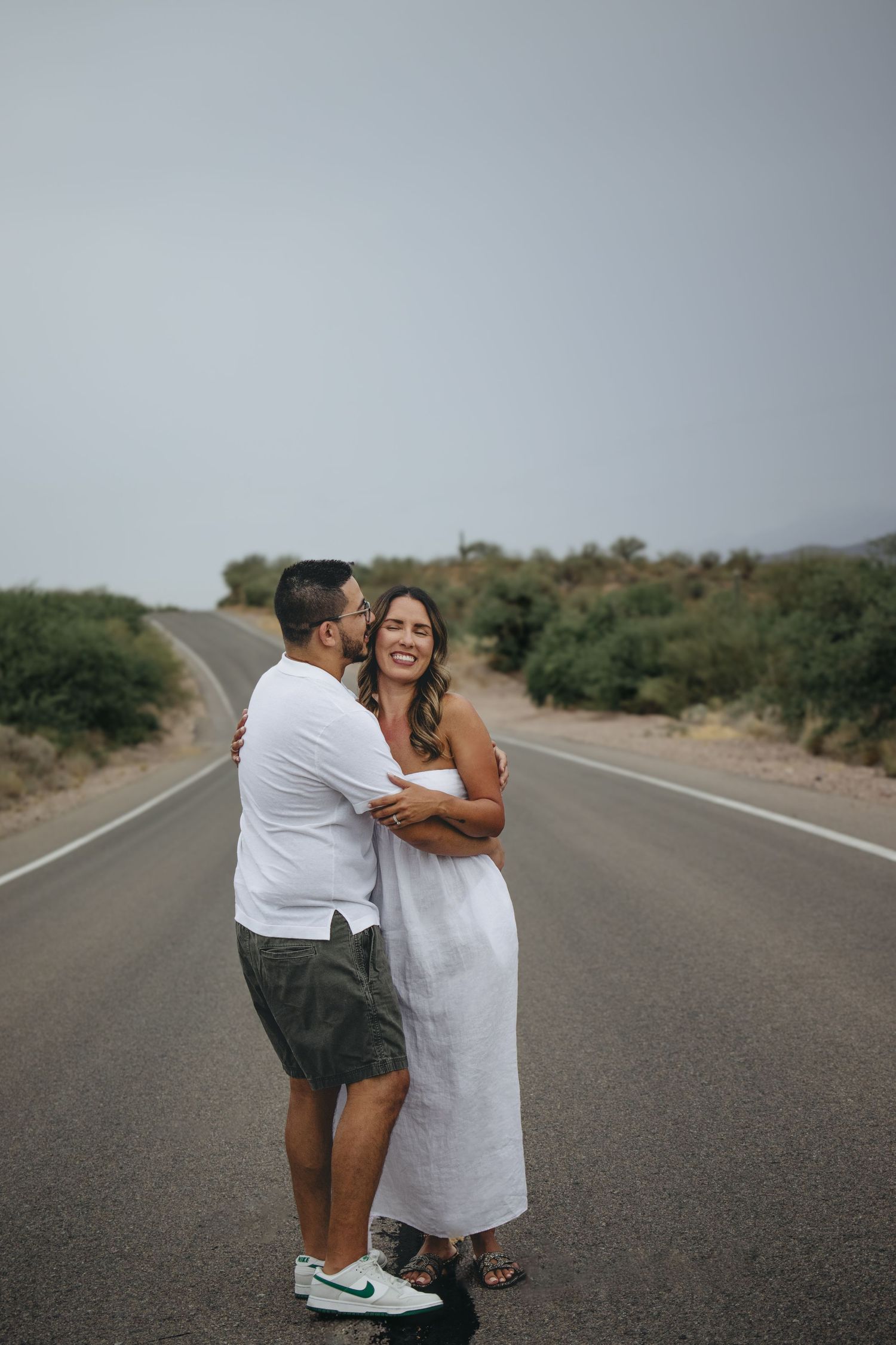 Couple hugging on a desert road, woman in white dress smiling as man embraces her from behind.