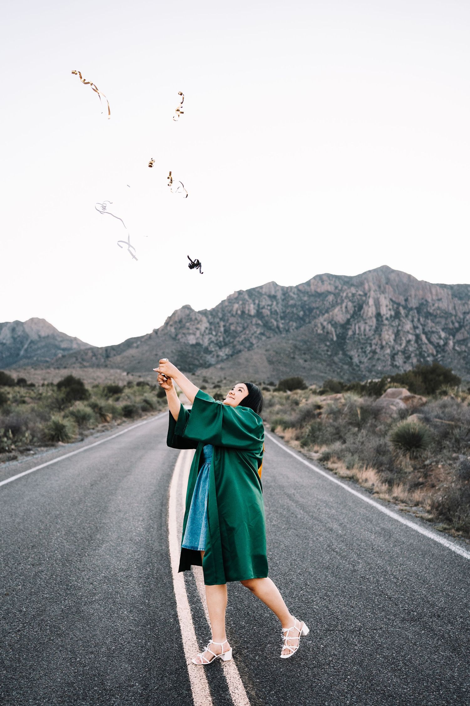 Graduate in green gown tossing items in air on empty desert road with mountain backdrop.