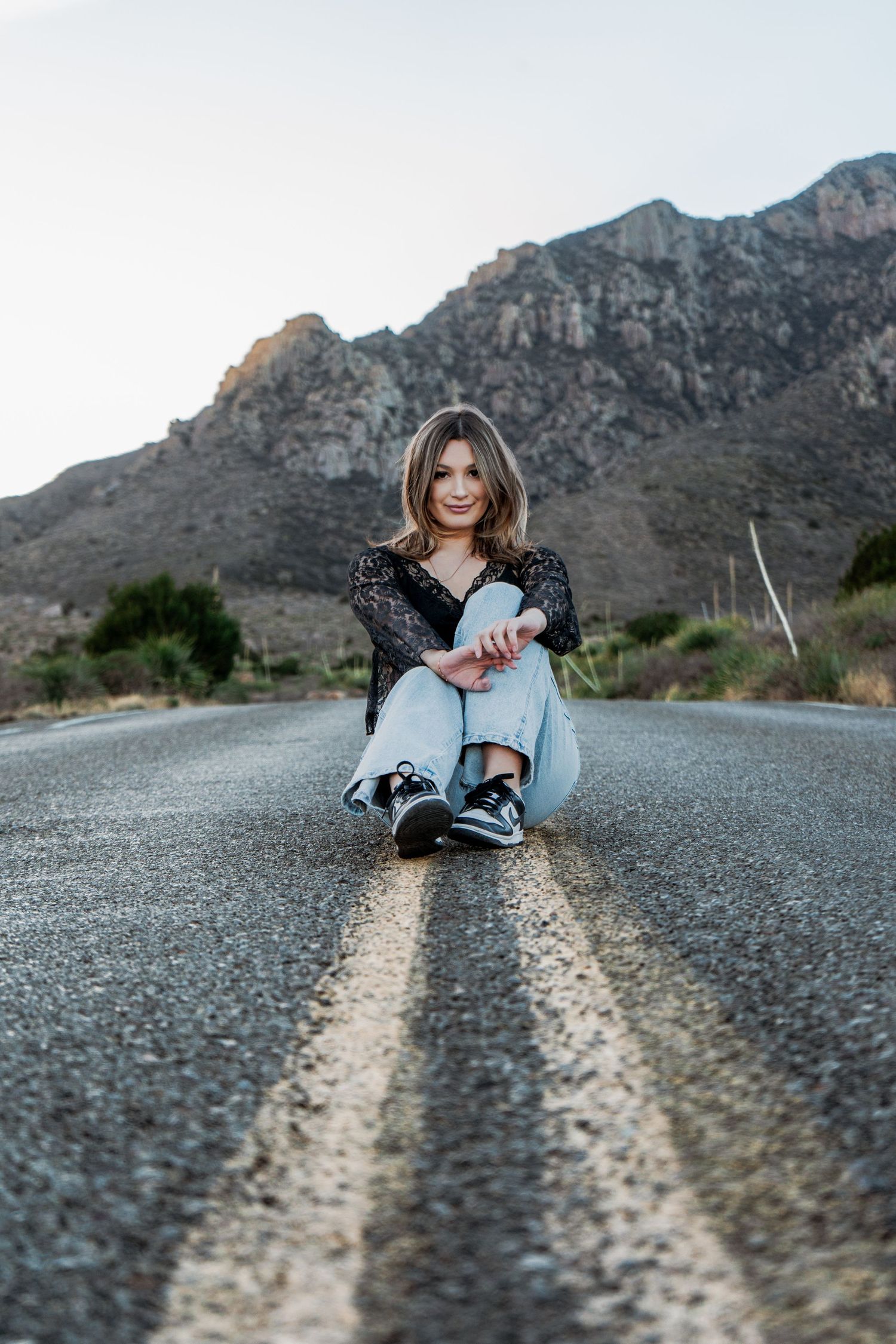 Young woman sitting on road centerline with mountain backdrop in casual jeans and black lace top.