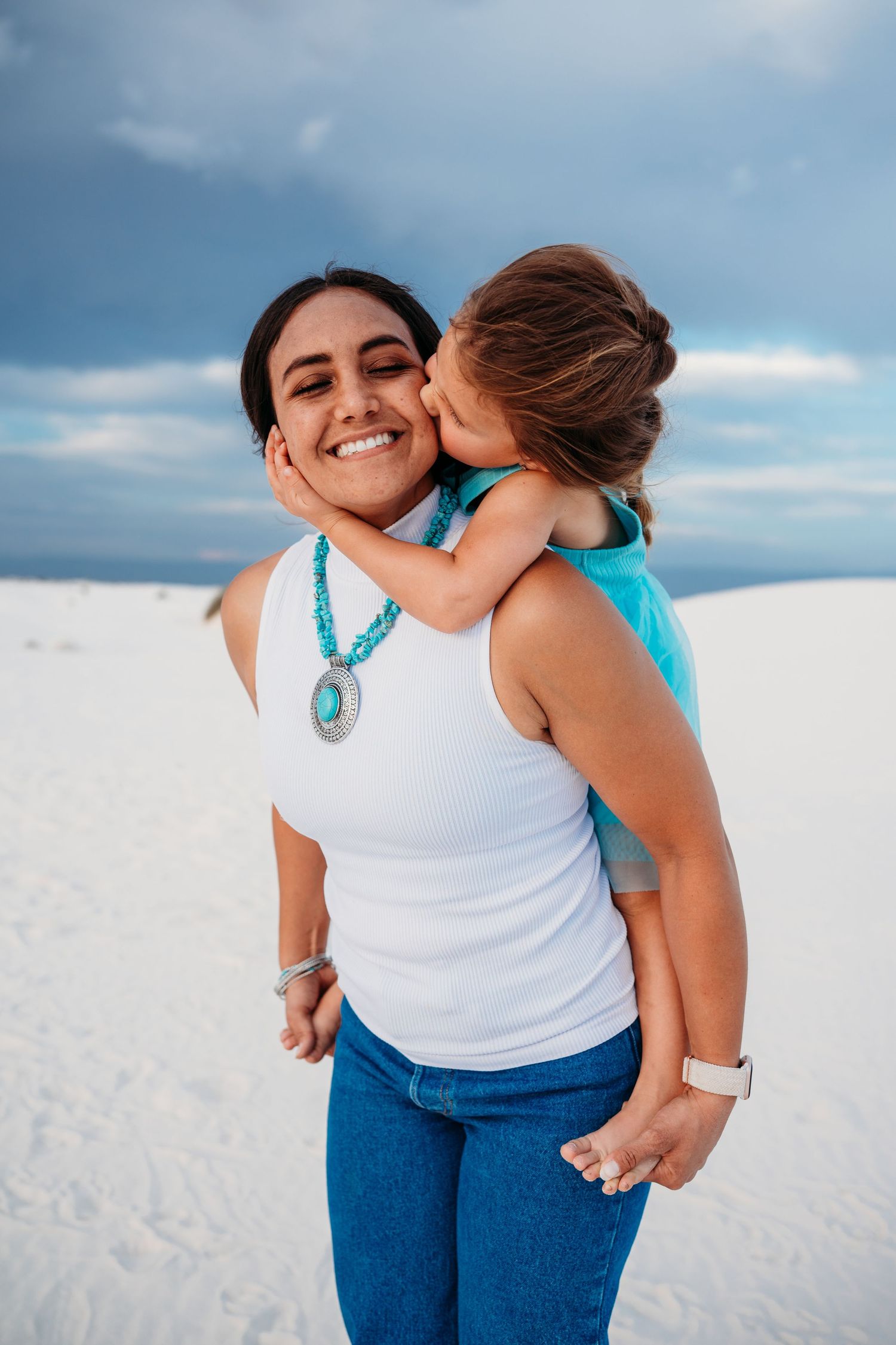 Young girl in turquoise dress kissing smiling mother wearing turquoise necklace on white sand beach.
