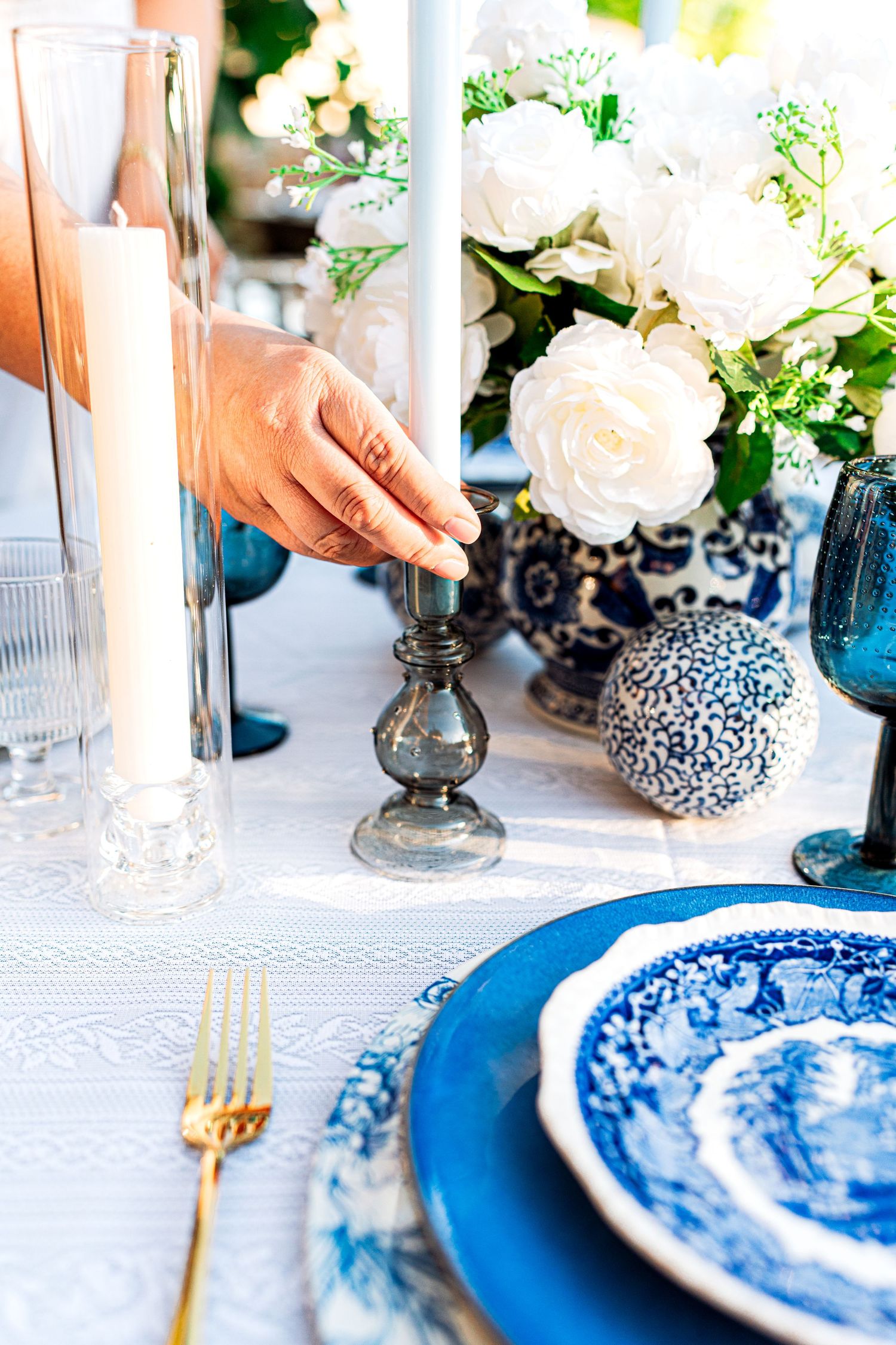 Hand adjusting a silver candlestick on an elegant blue and white outdoor tablescape with white roses.