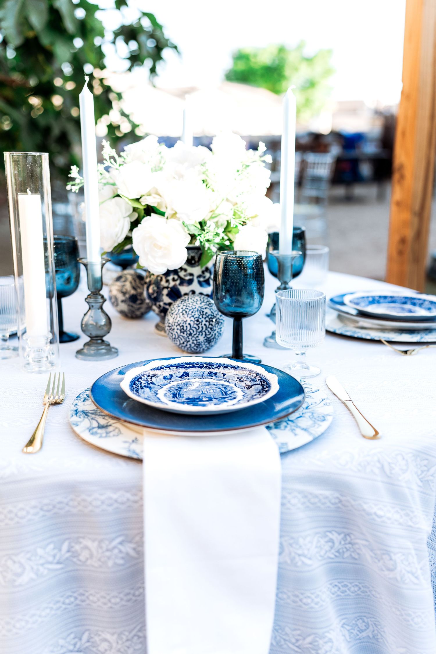 Sophisticated blue and white table setting with gold flatware, white napkin, and candlestick centerpieces outdoors.
