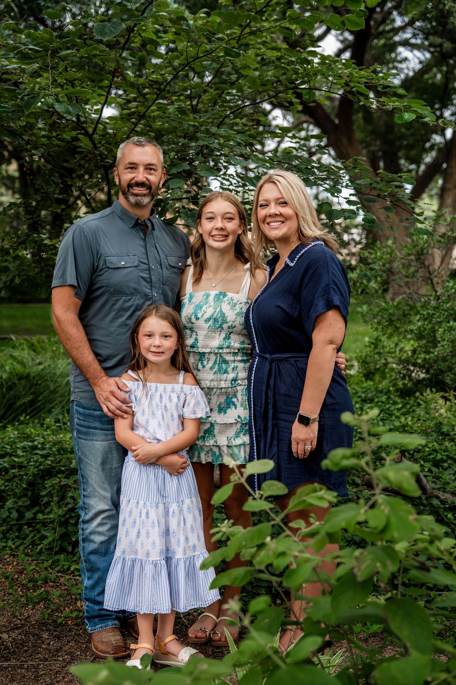 Family of four smiling together surrounded by lush green foliage in garden setting.