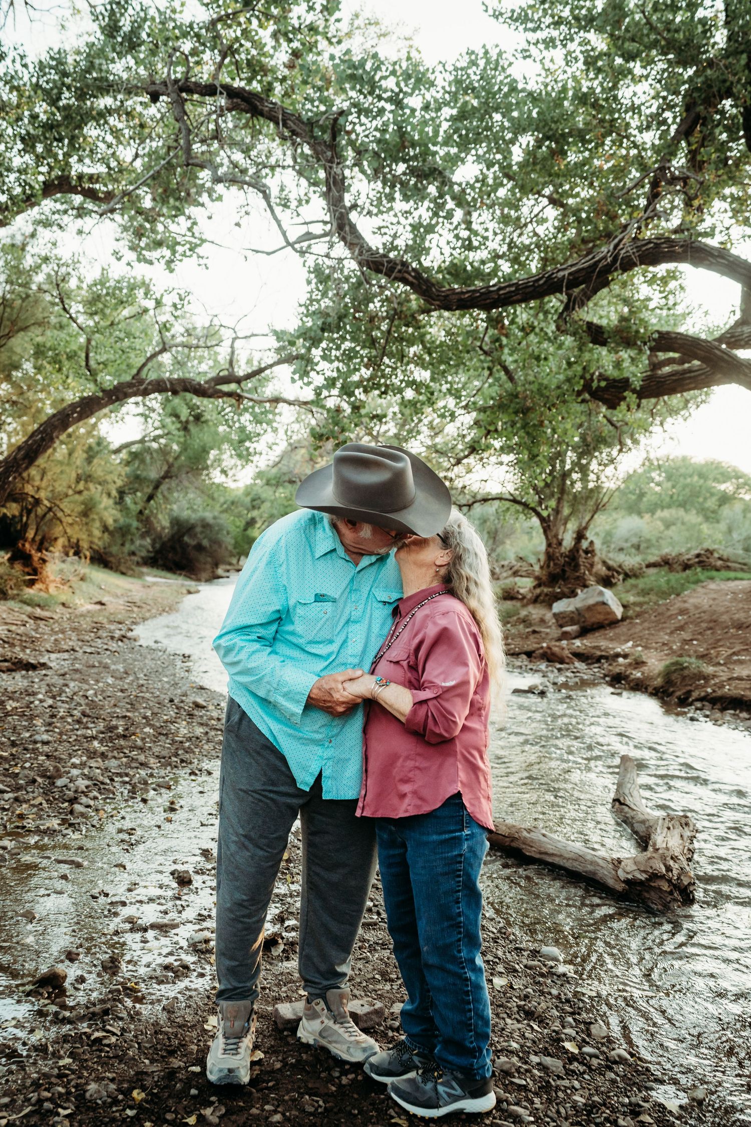 An older couple shares a romantic kiss beside a peaceful rocky creek shaded by large trees.