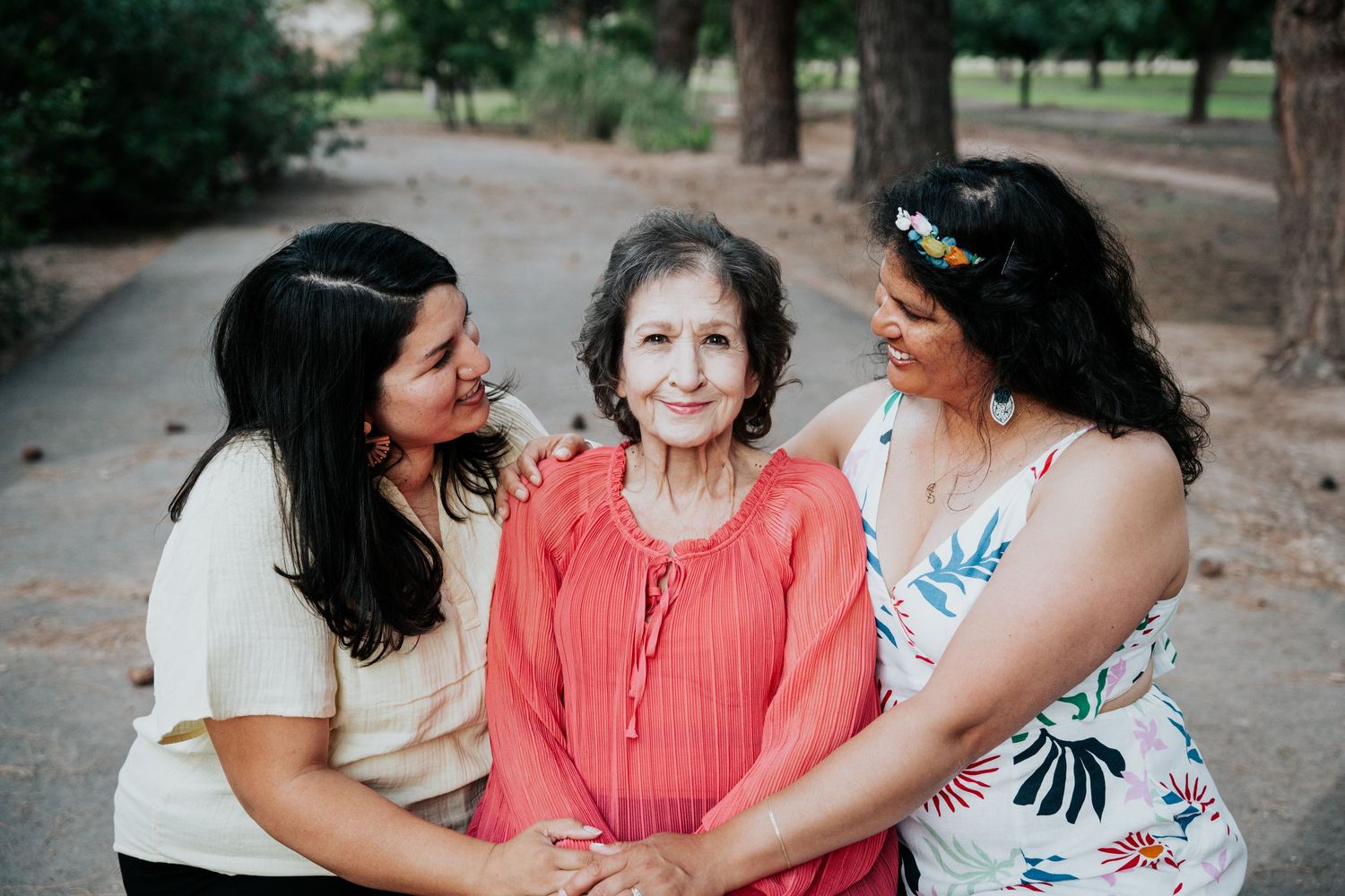 Three women share a tender moment together on a tree-lined path, the eldest in the center.
