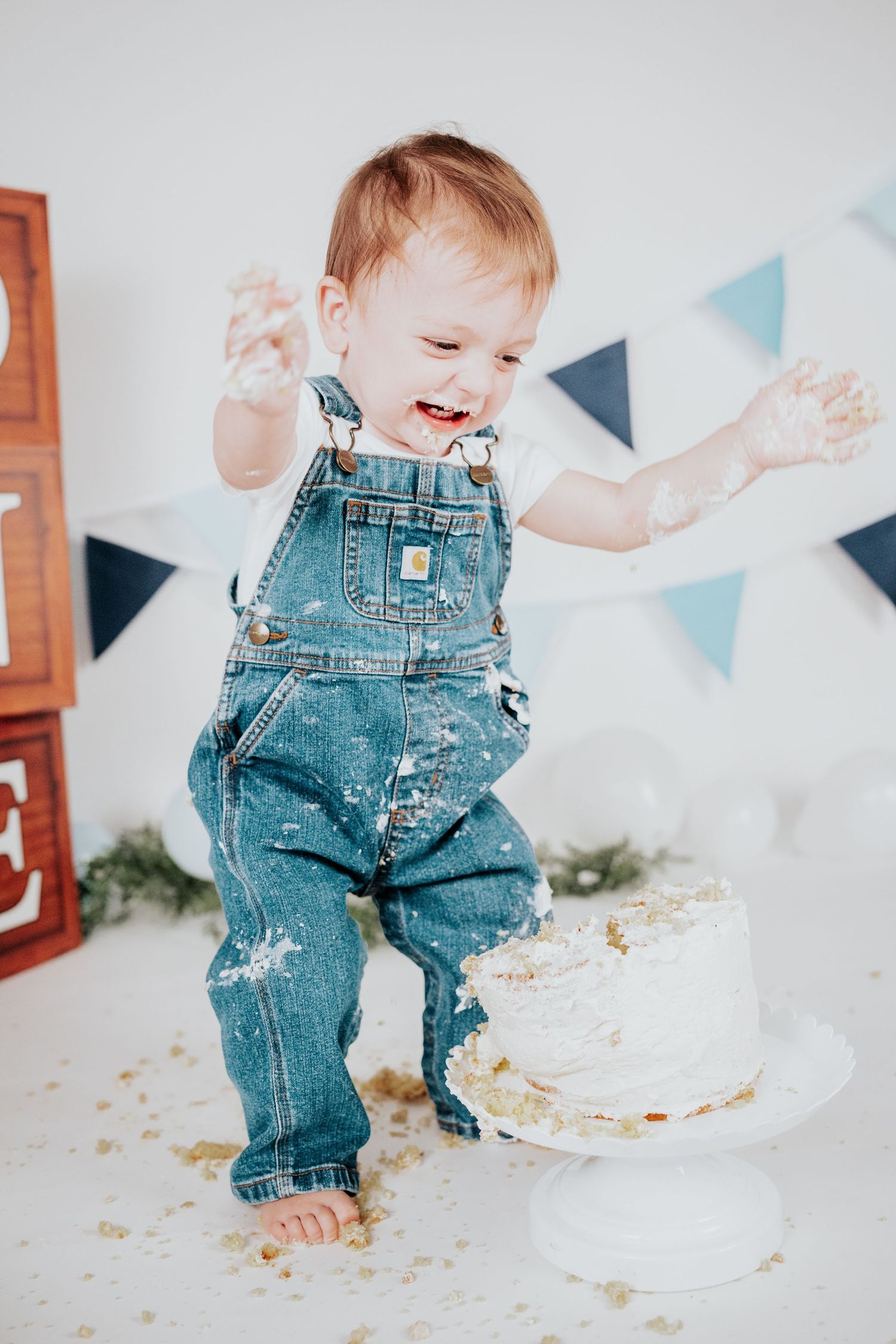 Laughing baby boy in denim overalls celebrating first birthday cake smash with frosting on hands.