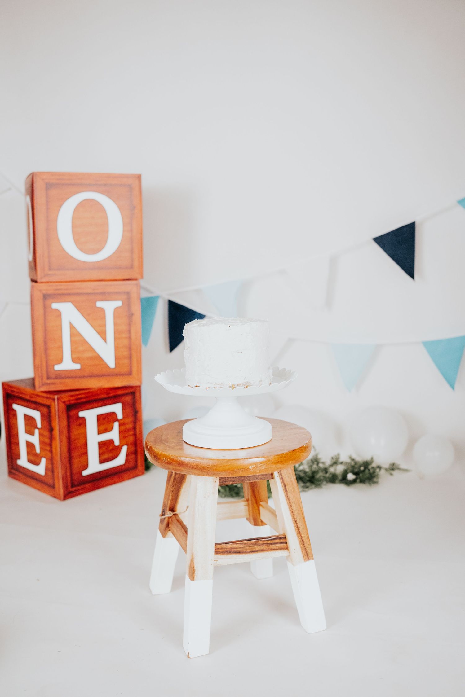 First birthday cake smash setup with wooden ONE blocks, white cake on stool, and blue bunting decor.