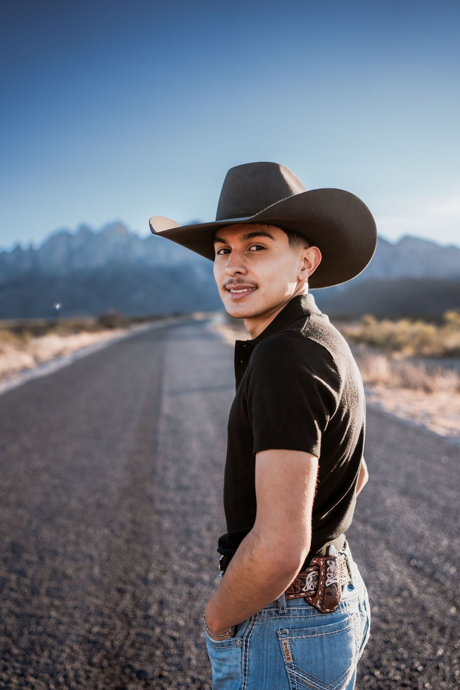 Young man in black cowboy hat and jeans poses on open desert road with mountains in background.