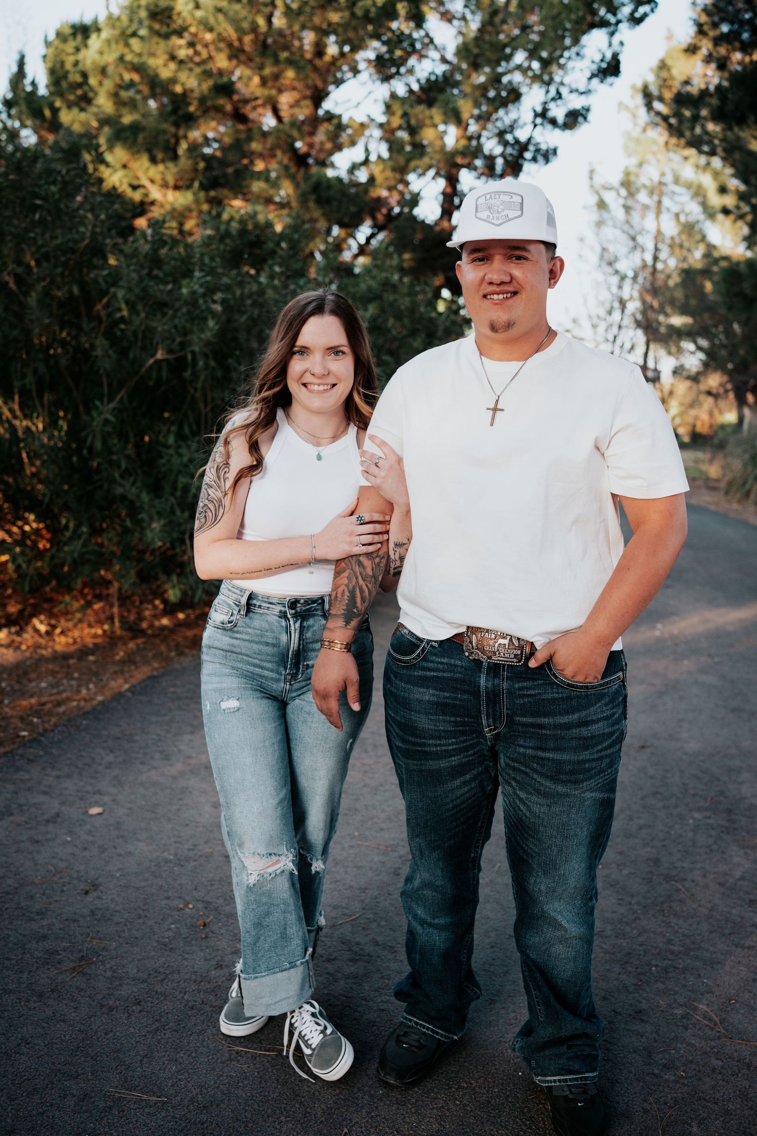 Smiling couple in matching white shirts and jeans posing on a tree-lined path in golden hour light.