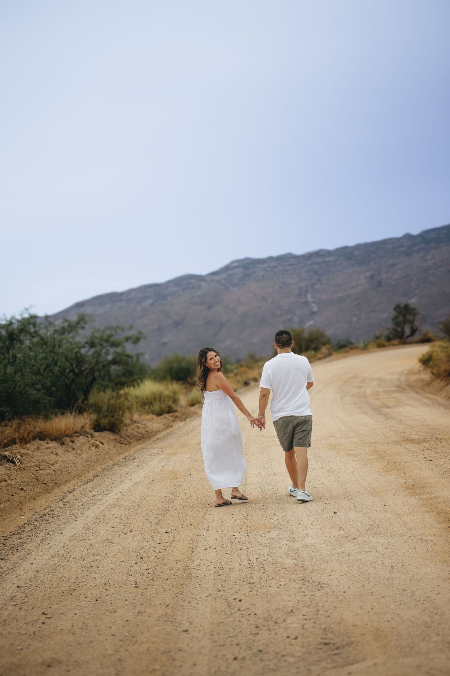 Couple holding hands walking down a dusty desert road with mountains in the background.